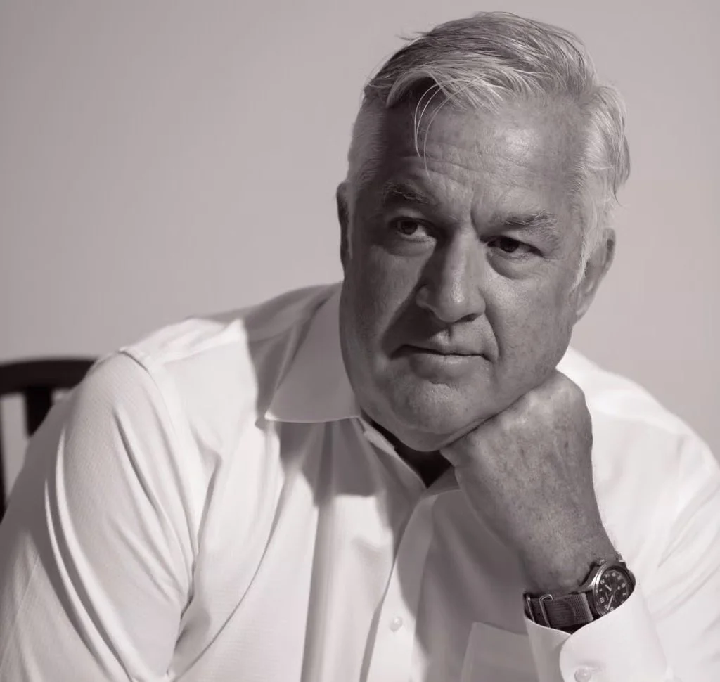 Black and white portrait of an older man with gray hair, wearing a white shirt, resting his chin on his hand, and wearing a watch.