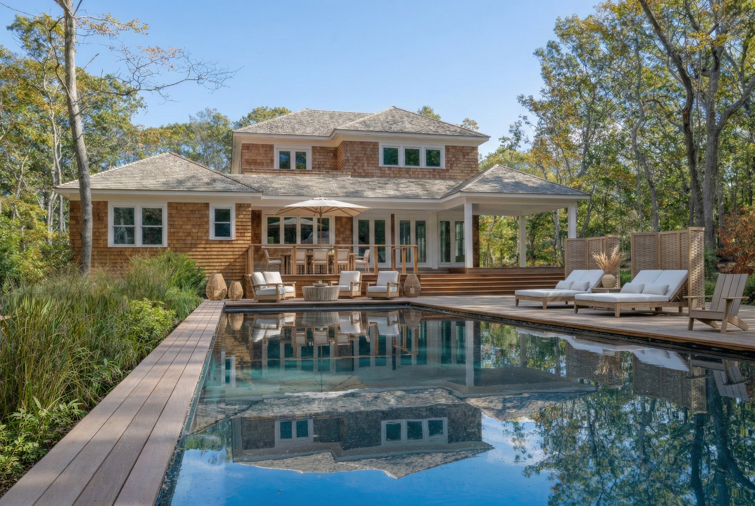 Modern house with wooden exterior and large windows, surrounded by trees, with a swimming pool in the foreground and outdoor furniture on the deck.