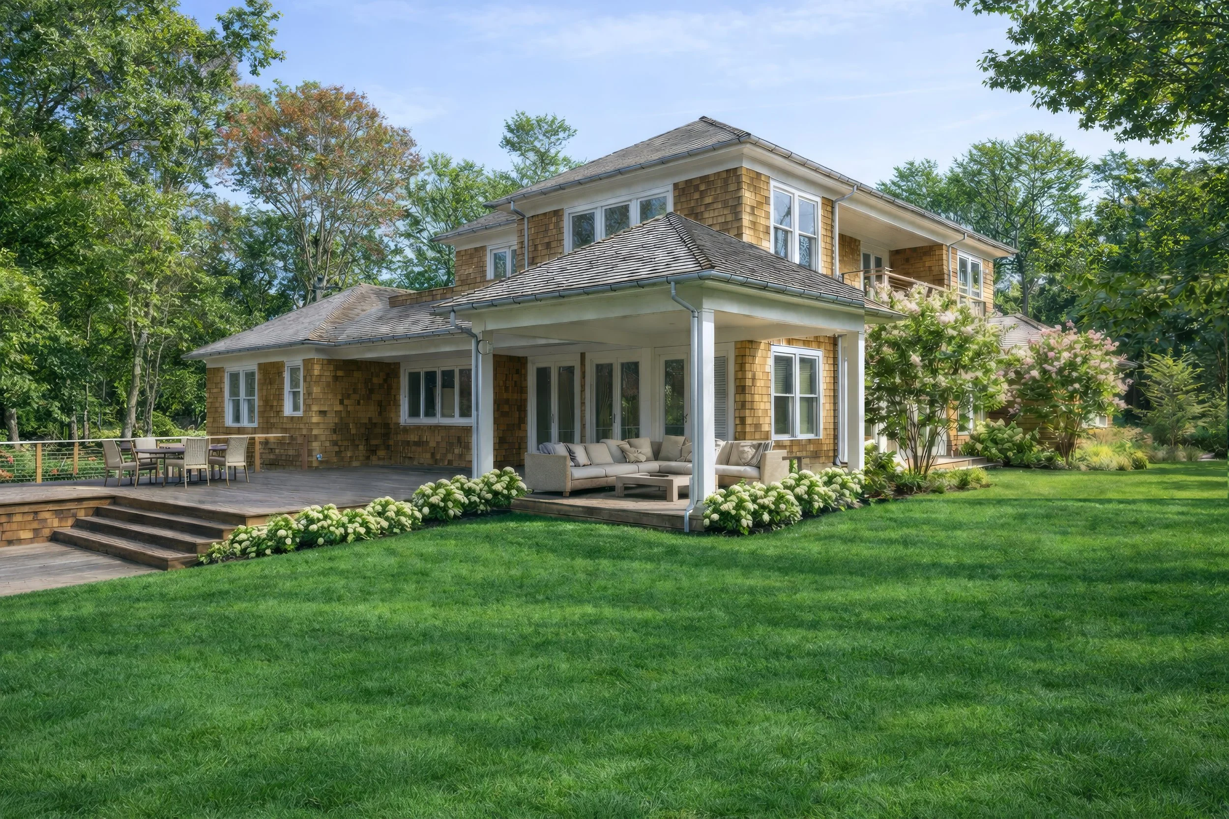 A large two-story house with wooden shingle siding, white trim, and a covered porch with outdoor seating. The house is surrounded by a well-maintained lawn, blooming bushes, and tall trees, with a bright blue sky in the background.