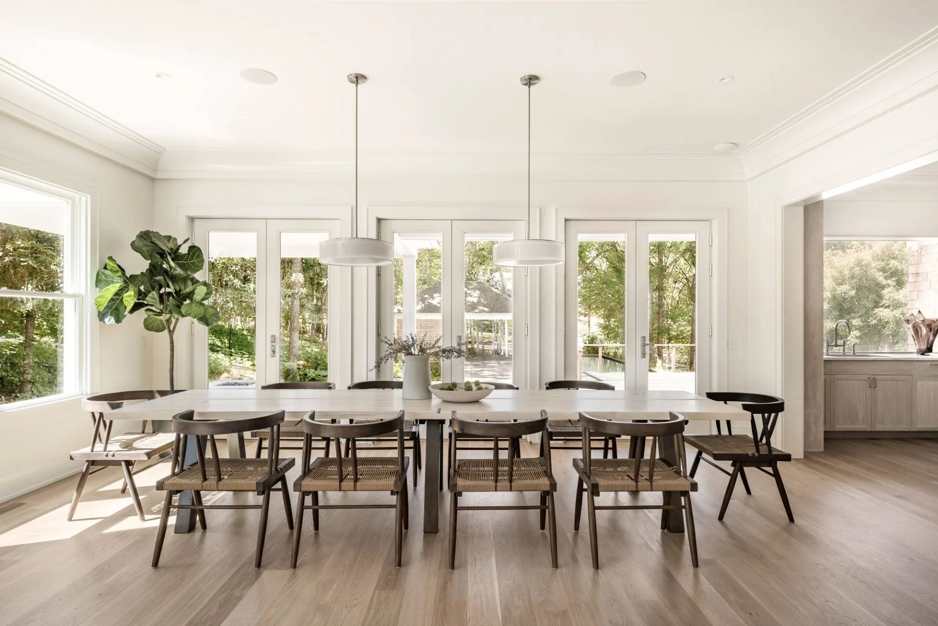 Bright dining room with a long white table, surrounded by wooden chairs with woven seats, large windows and glass doors with greenery outside, pendant lights overhead, and an open view to the kitchen area.