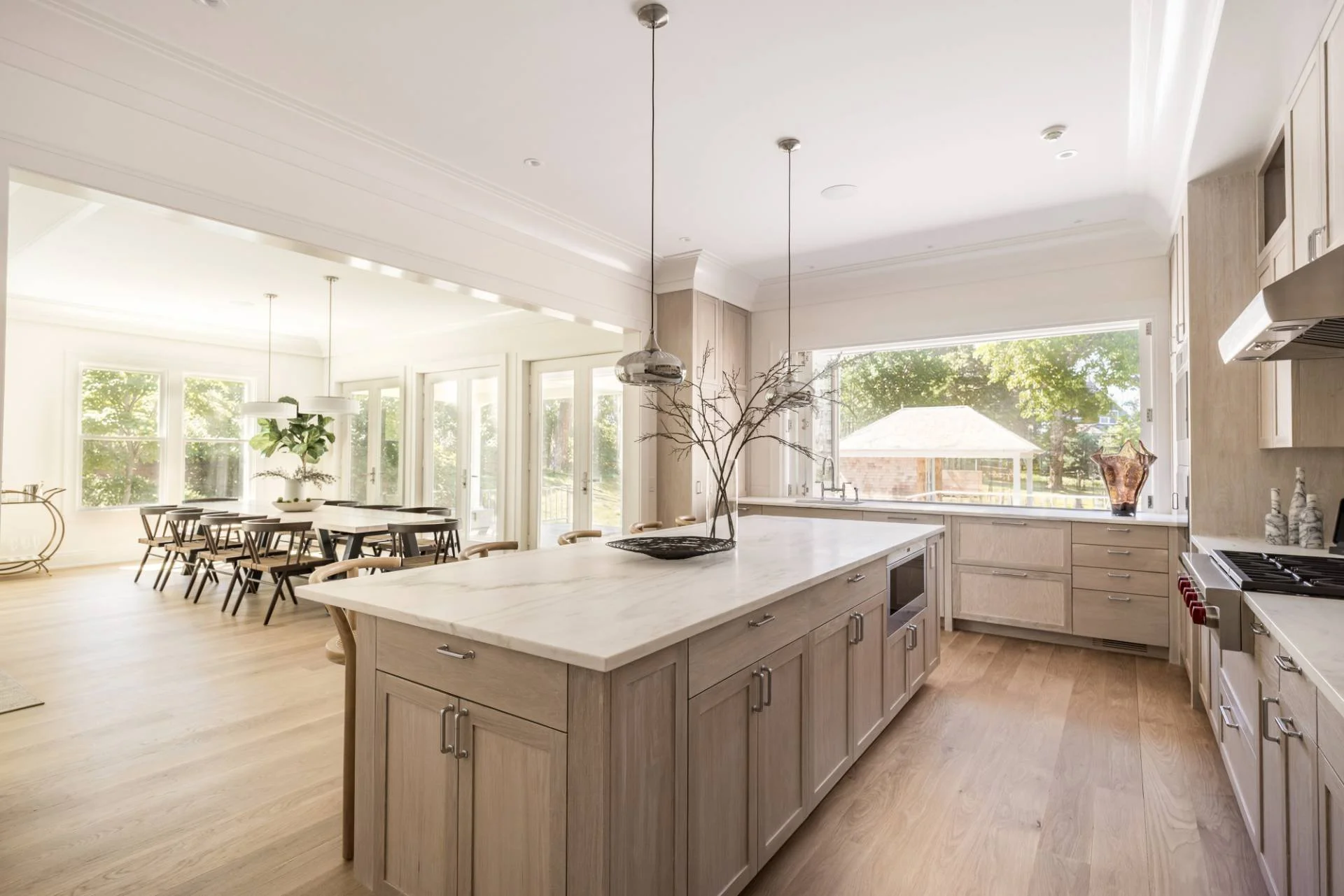 Bright, modern kitchen with a large island, light wood cabinets, and large windows overlooking a backyard. A bare tree branch centerpiece decorates the island, and a glass vessel is placed on the counter near the window. The adjacent dining area has 