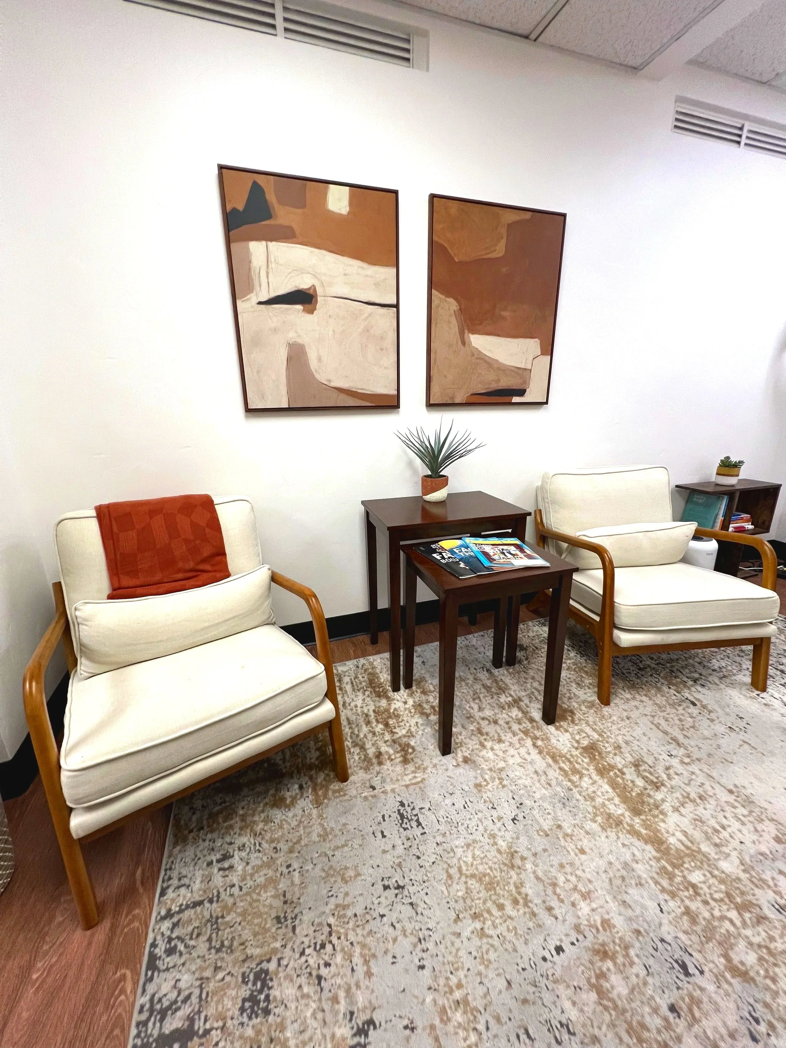 A cozy waiting area with two white armchairs with wooden frames, a small dark wood table with magazines, a potted plant, a second smaller table with books, and two abstract art paintings on the white wall behind.
