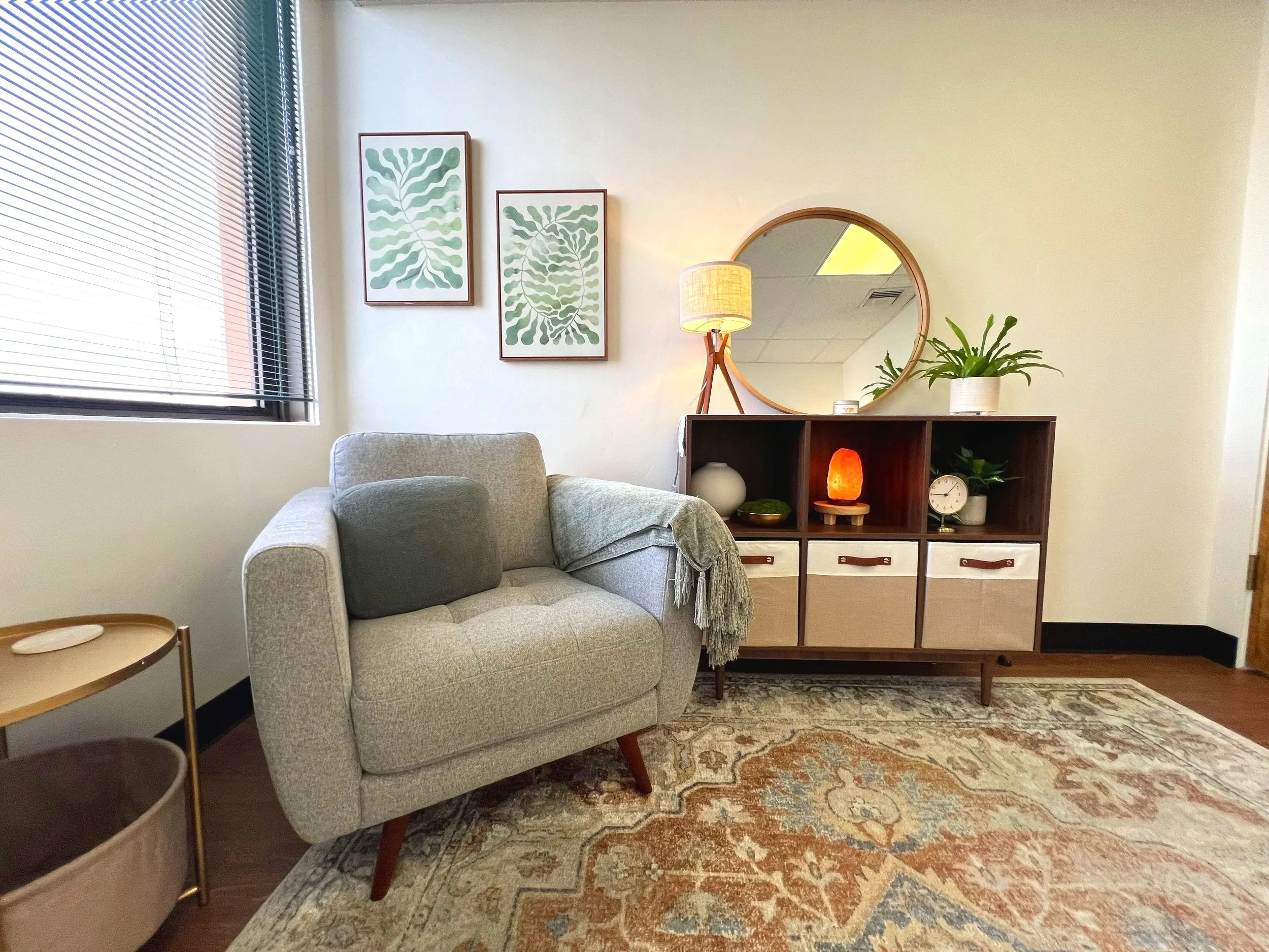 Therapy office with a gray armchair, a wooden side table, a patterned area rug, a sideboard with decorative items including a salt lamp, small vases, and plants, a round mirror mounted on the wall, and framed green botanical art on the wall.