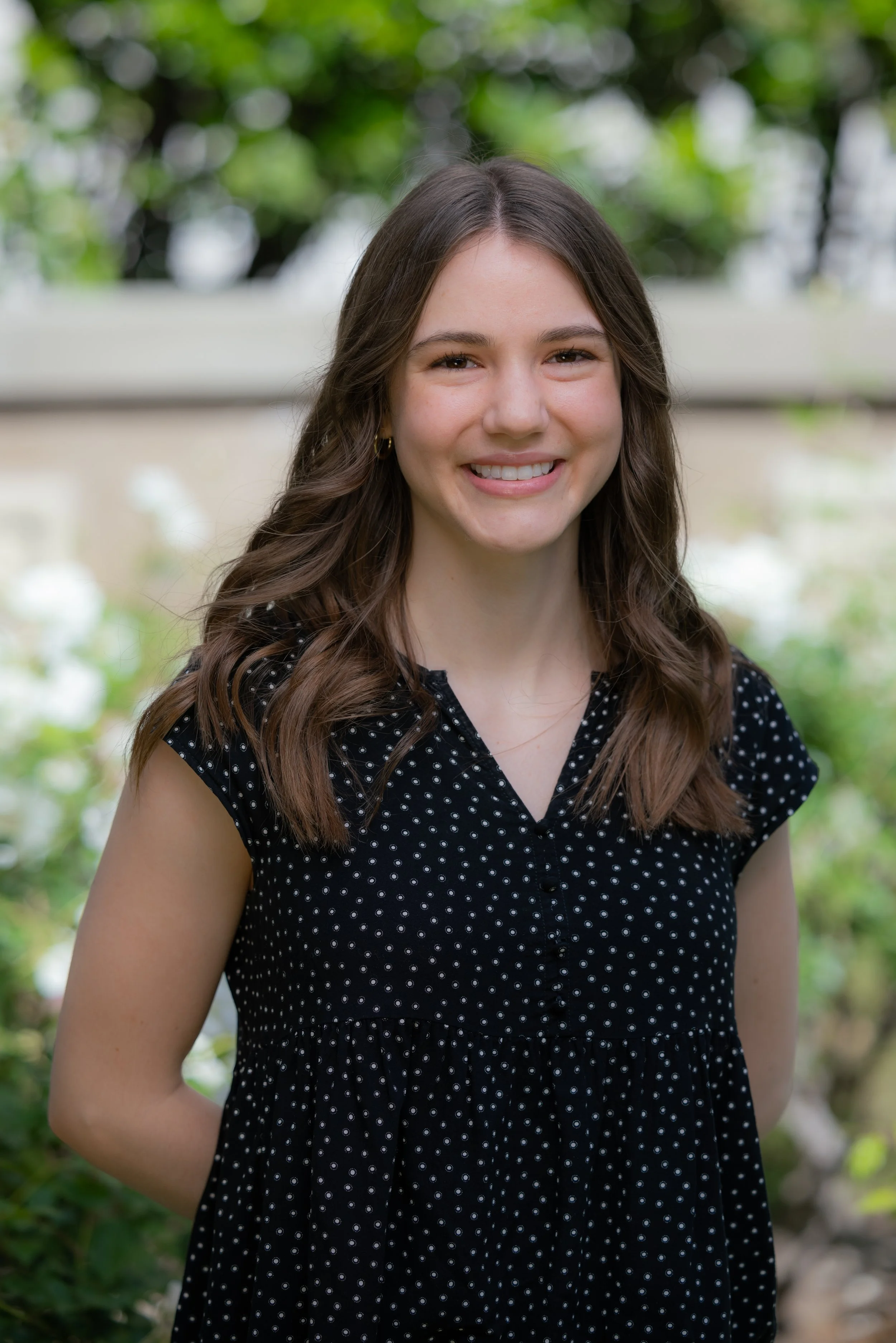 A young woman with long brown hair, smiling, wearing a black dress with small white polka dots, standing outdoors with greenery in the background.