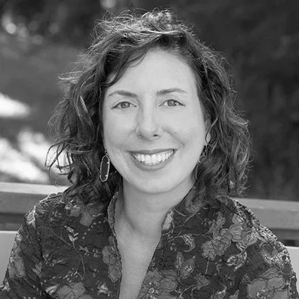 Black and white portrait of a woman with curly hair smiling, wearing earrings and a floral top, outdoors setting.