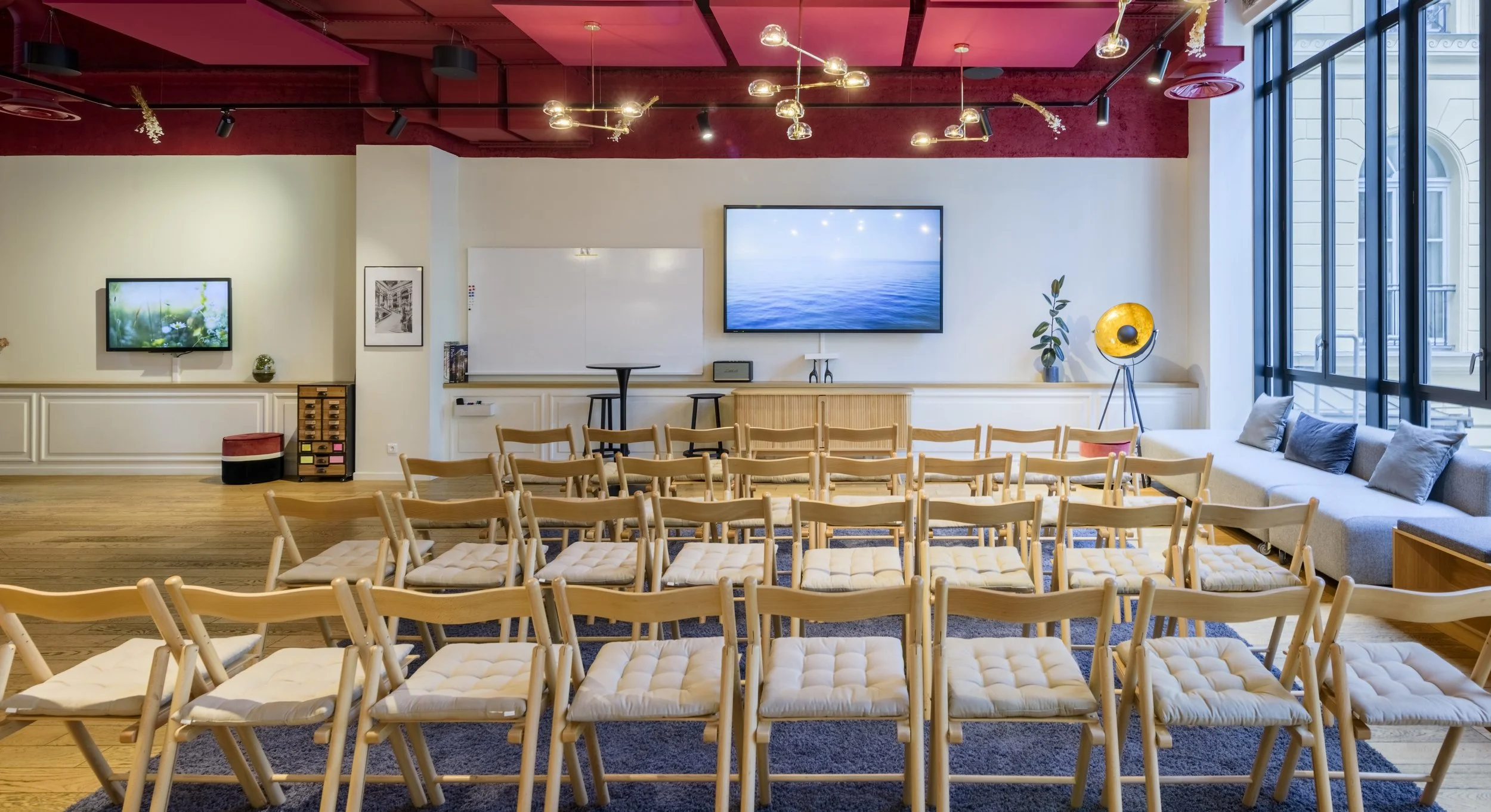 Salle de conférence avec chaises en bois, écran de projection et grandes fenêtres laissant entrer la lumière