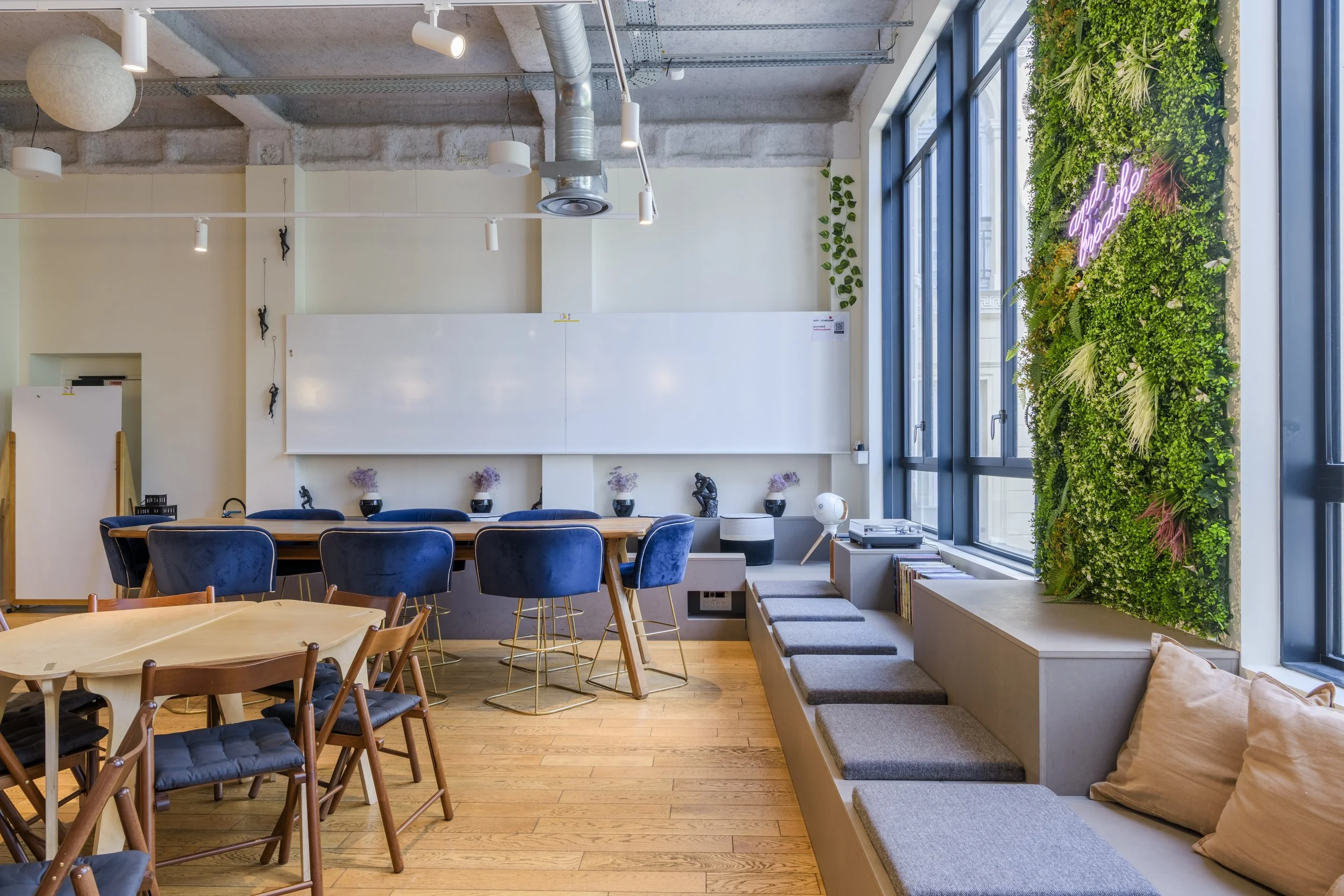 Intérieur d'un espace de coworking lumineux avec des murs en verre, un mur végétal avec une enseigne néon rose, une table en bois avec des chaises bleues, et un coin salon avec des coussins.