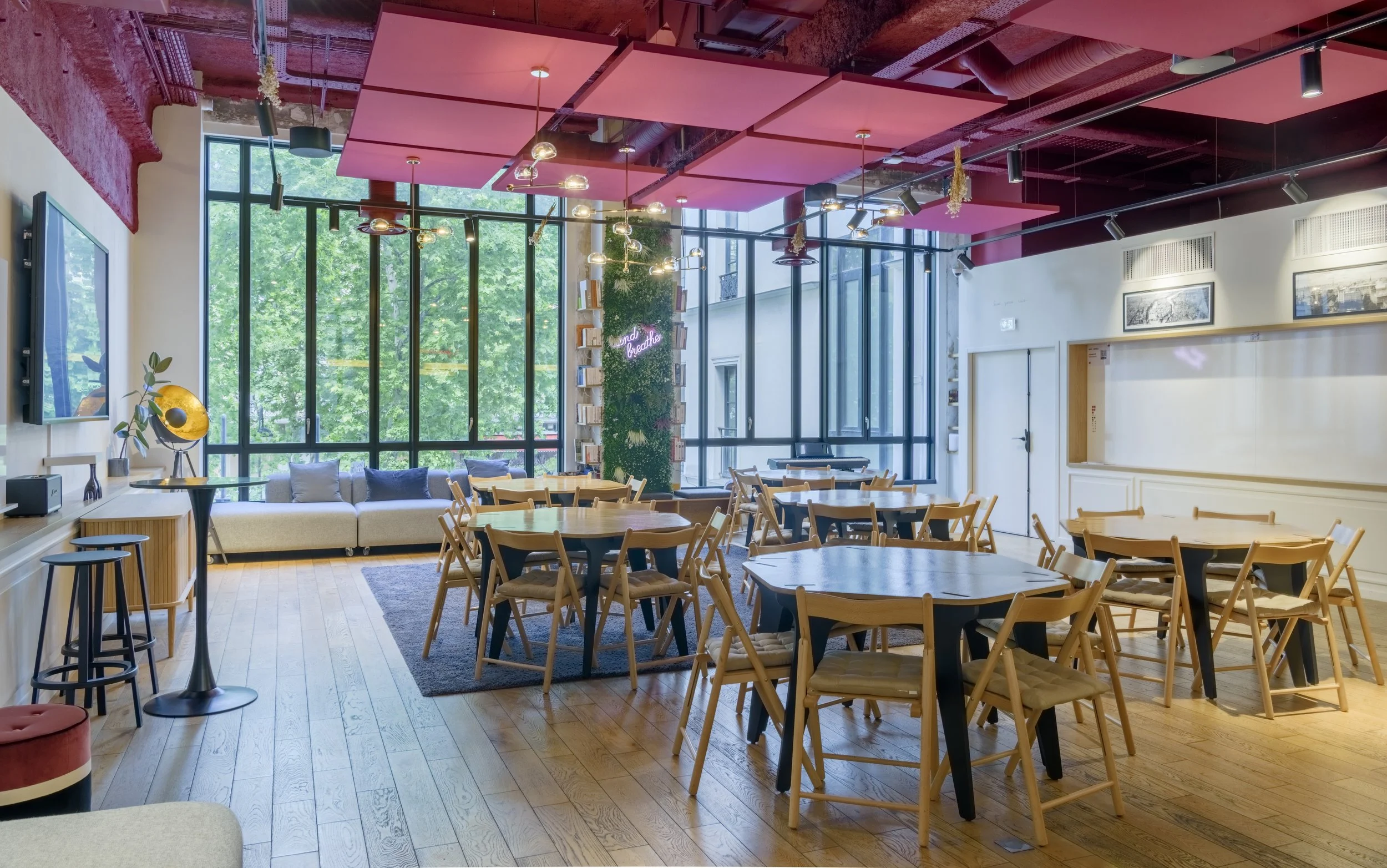 Intérieur d'un café ou restaurant moderne, avec plusieurs tables en bois, des chaises en bois clair, un canapé beige près de grandes fenêtres laissant entrer beaucoup de lumière naturelle, un mur décoratif avec des étagères et un néon rose, et un plafond avec des panneaux roses suspendus.