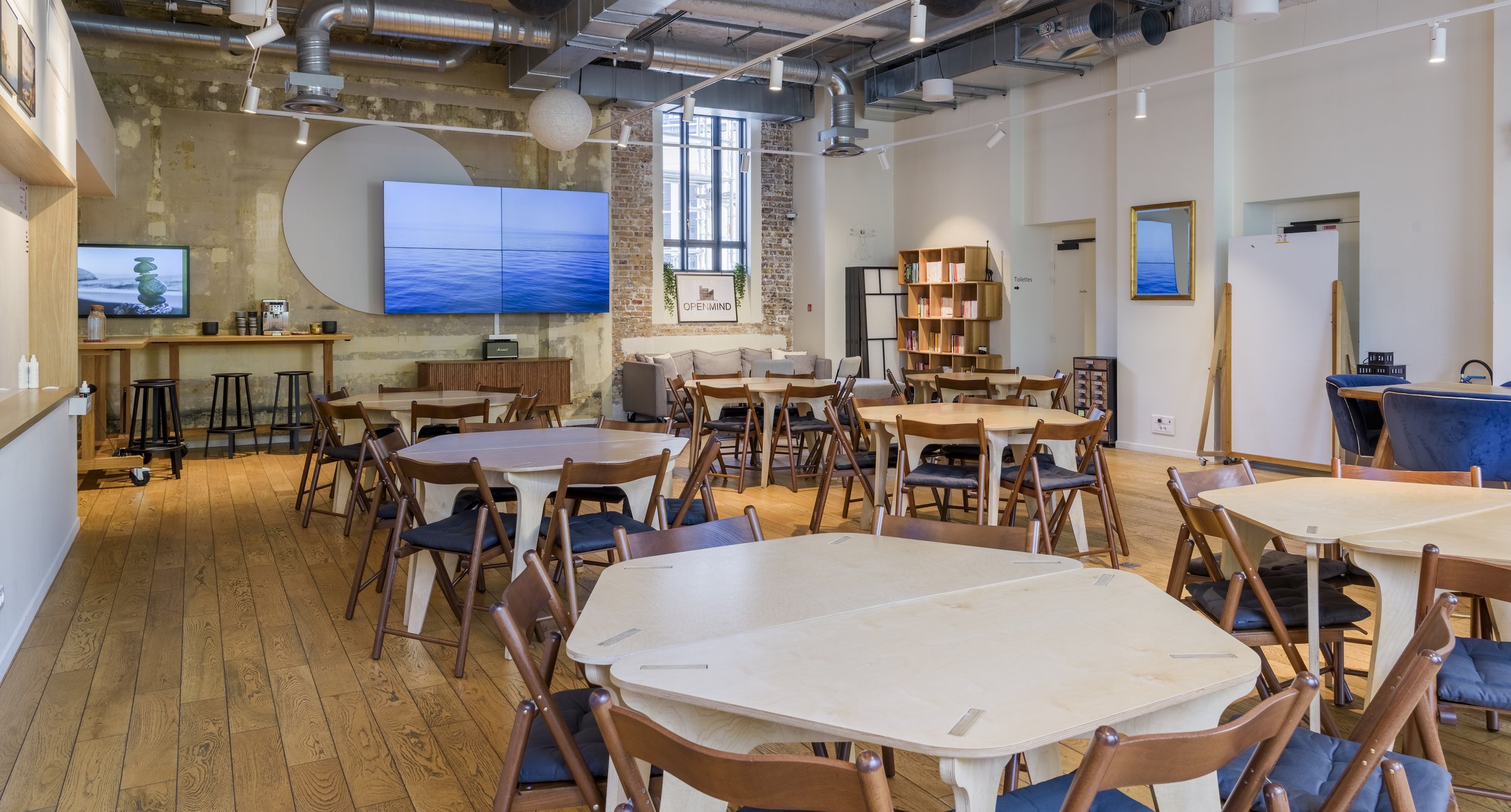 Salle de réunion ou espace de coworking avec des tables en bois, des chaises en bois avec coussins noirs, une bibliothèque, des écrans, et un mur en briques dans un environnement moderne et lumineux.