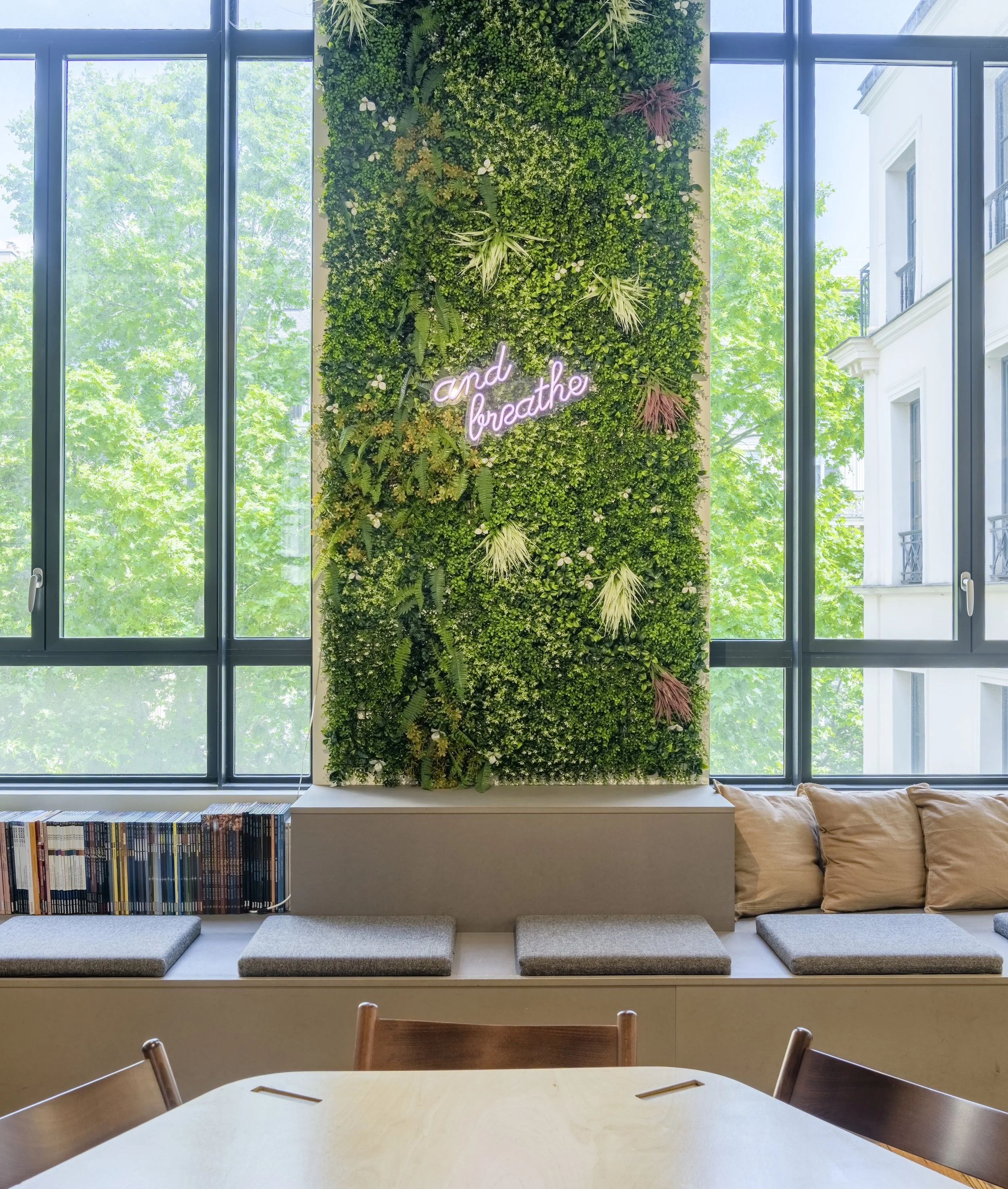 Mur végétal avec une pancarte lumineuse disant « and breathe » dans un intérieur lumineux avec de grandes fenêtres, des coussins et des livres.