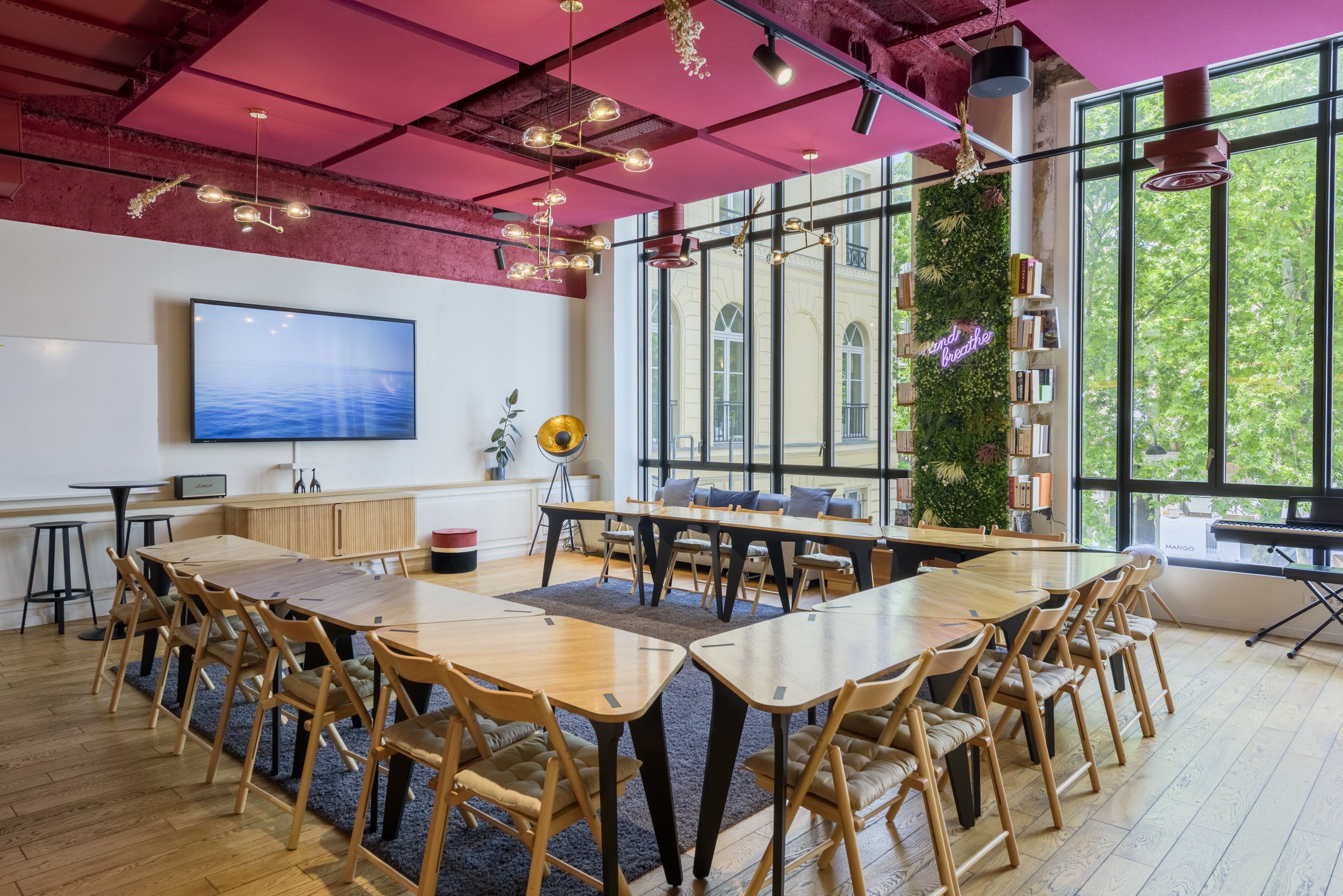 Salle de réunion moderne avec tables en bois, chaises, grand mur de fenêtres, décoration verte, néons roses et suspensions lumineuses