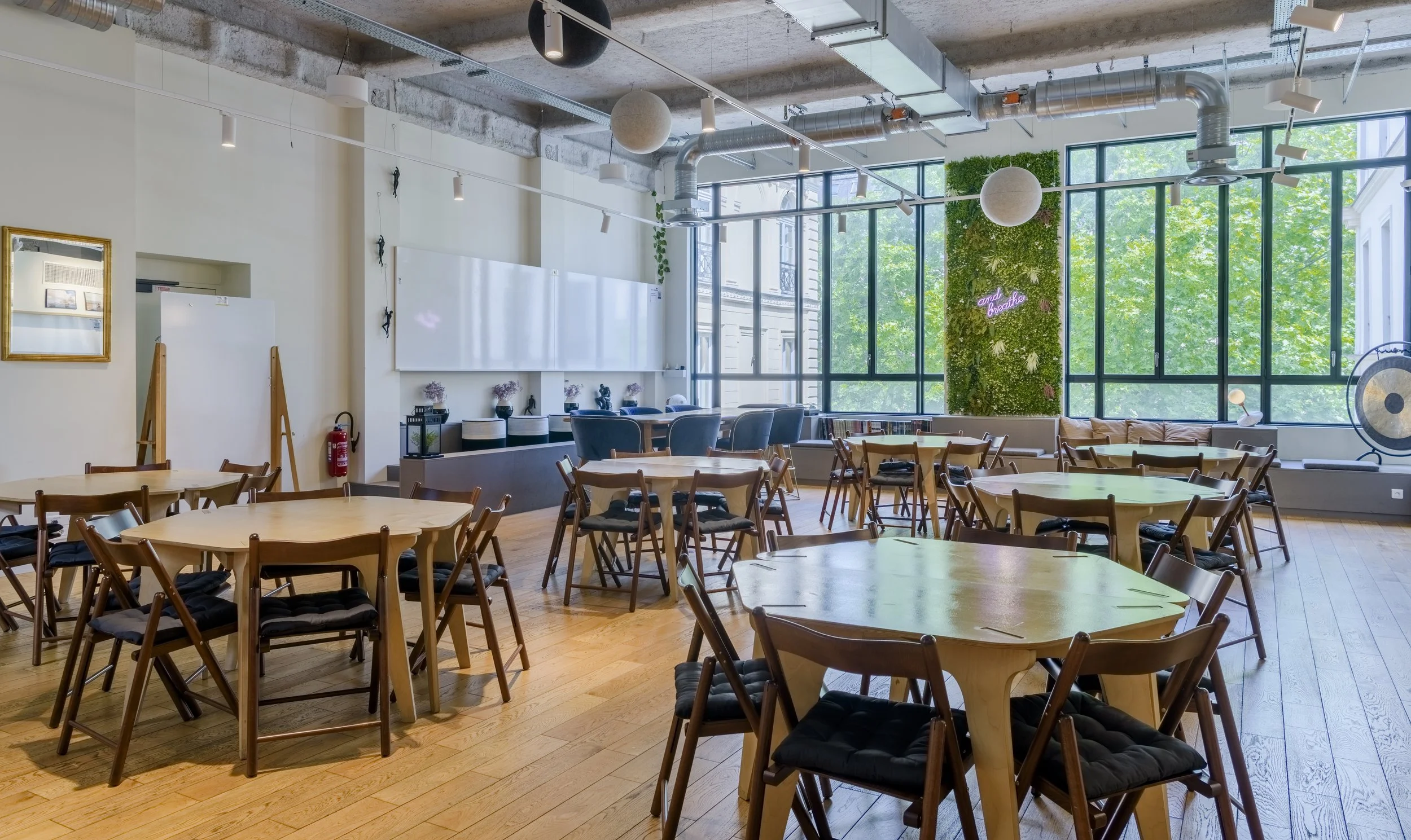 Salle de réunion ou espace de coworking avec plusieurs tables rondes en bois, chaises en bois avec coussins noirs, grande baie vitrée laissant entrer la lumière naturelle, mur végétal avec une inscription néon rose 'and breathe', décoration minimalis