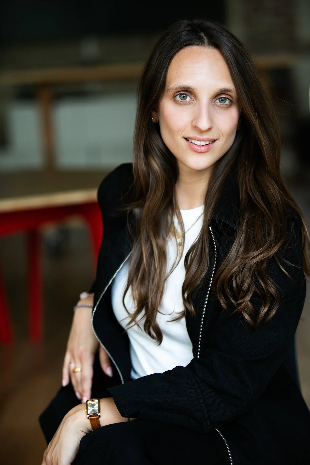 Jeune femme avec des cheveux longs, cheveux bruns, portant un manteau noir, assise dans une ambiance intérieure, souriante, regardant l'objectif.
