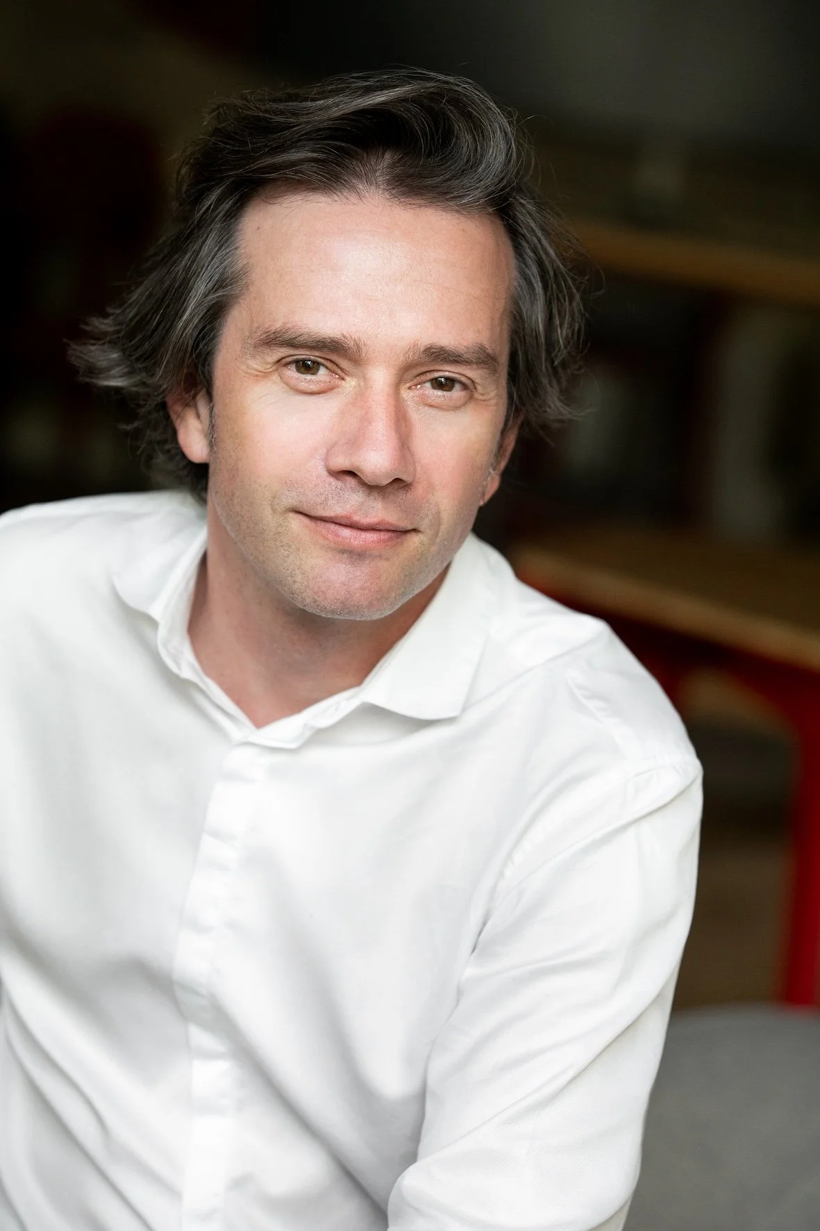 Hommes d'âge moyen avec cheveux bruns ondulés, portant une chemise blanche, souriant doucement, posé dans un intérieur avec des étagères en arrière-plan.