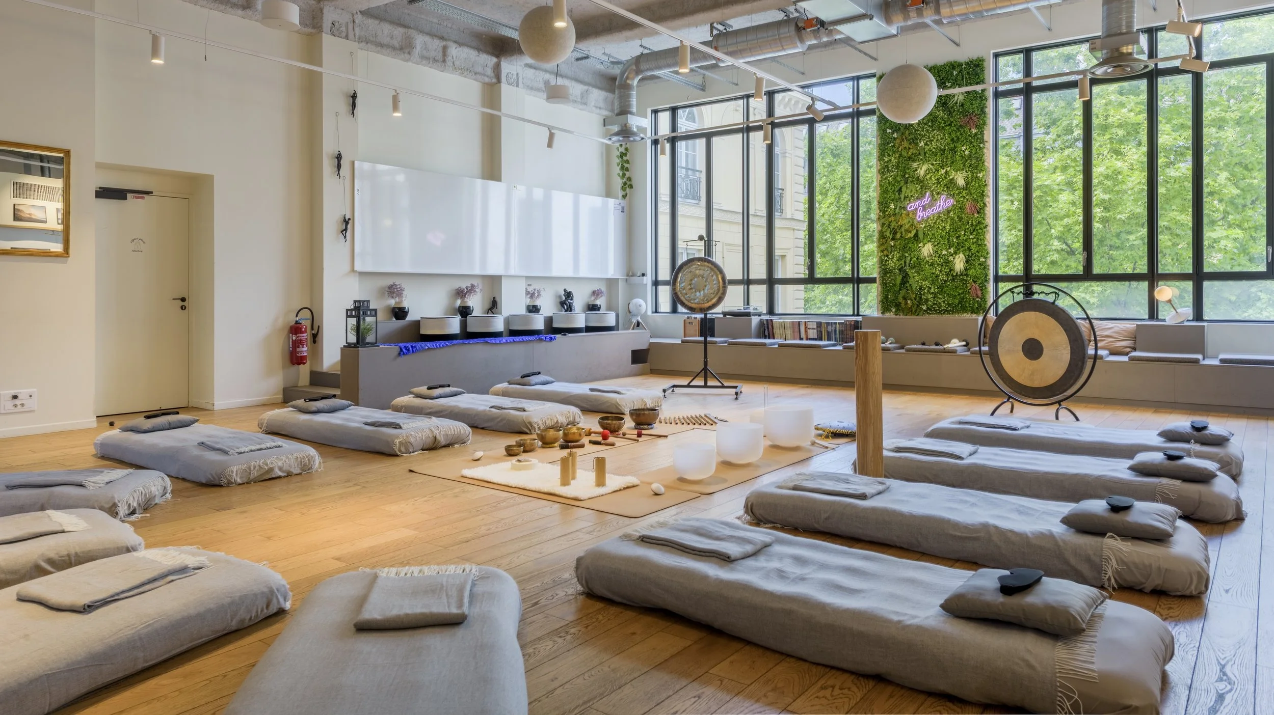 Salle de yoga ou méditation avec des tapis, coussins et instruments de musique dans une pièce lumineuse avec de grandes fenêtres et un mur végétal décoratif.