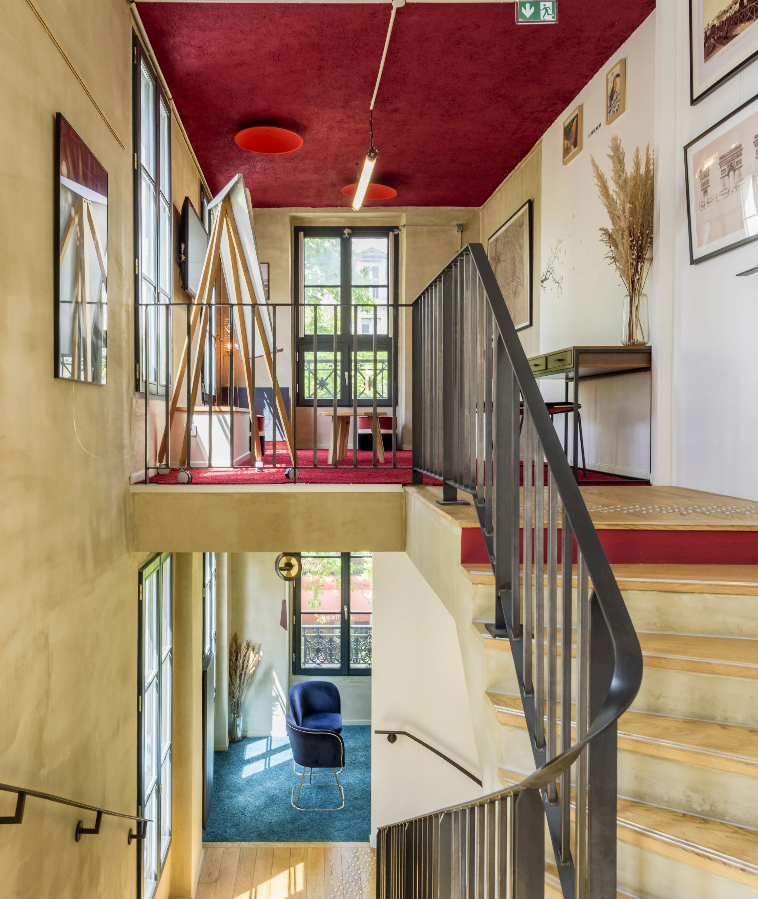 Intérieur lumineux avec escalier en colimaçon, murs jaunes, plafond rouge, grandes fenêtres, espace avec fauteuil bleu, œuvres d'art encadrées, plantes et mobilier moderne.