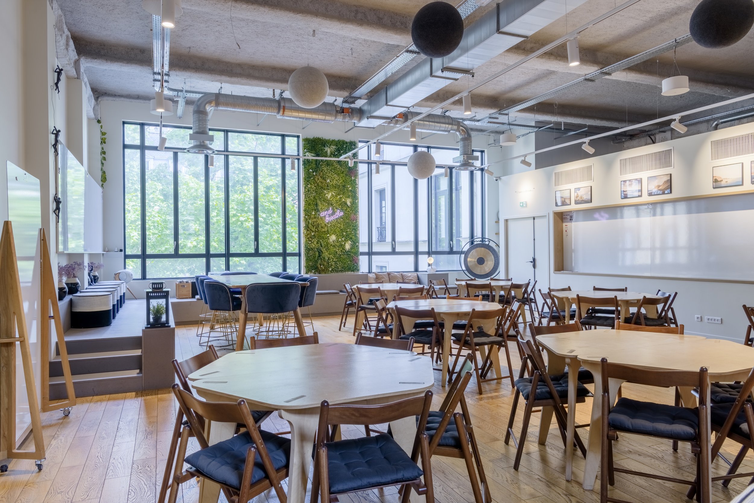 Une salle de réunion ou café avec des tables rondes en bois, chaises en bois et rembourrées, grandes fenêtres laissant entrer la lumière naturelle, décoration végétale, murs blancs avec des photos encadrées, un gong en arrière-plan et un mur vert ave