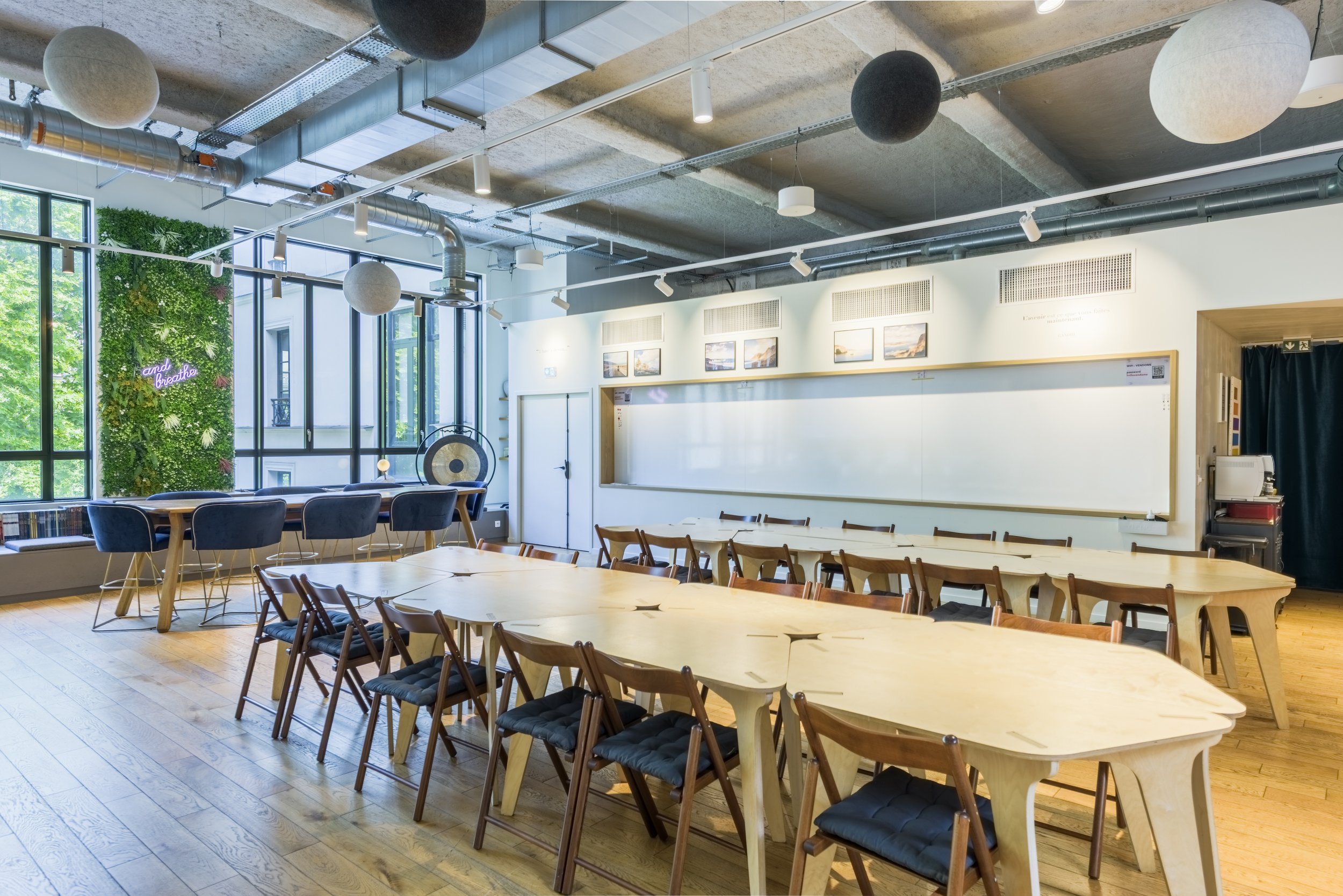 Salle de classe ou de réunion avec tables en bois, chaises, tableau blanc, décor moderne et fenêtres laissant entrer la lumière naturelle.