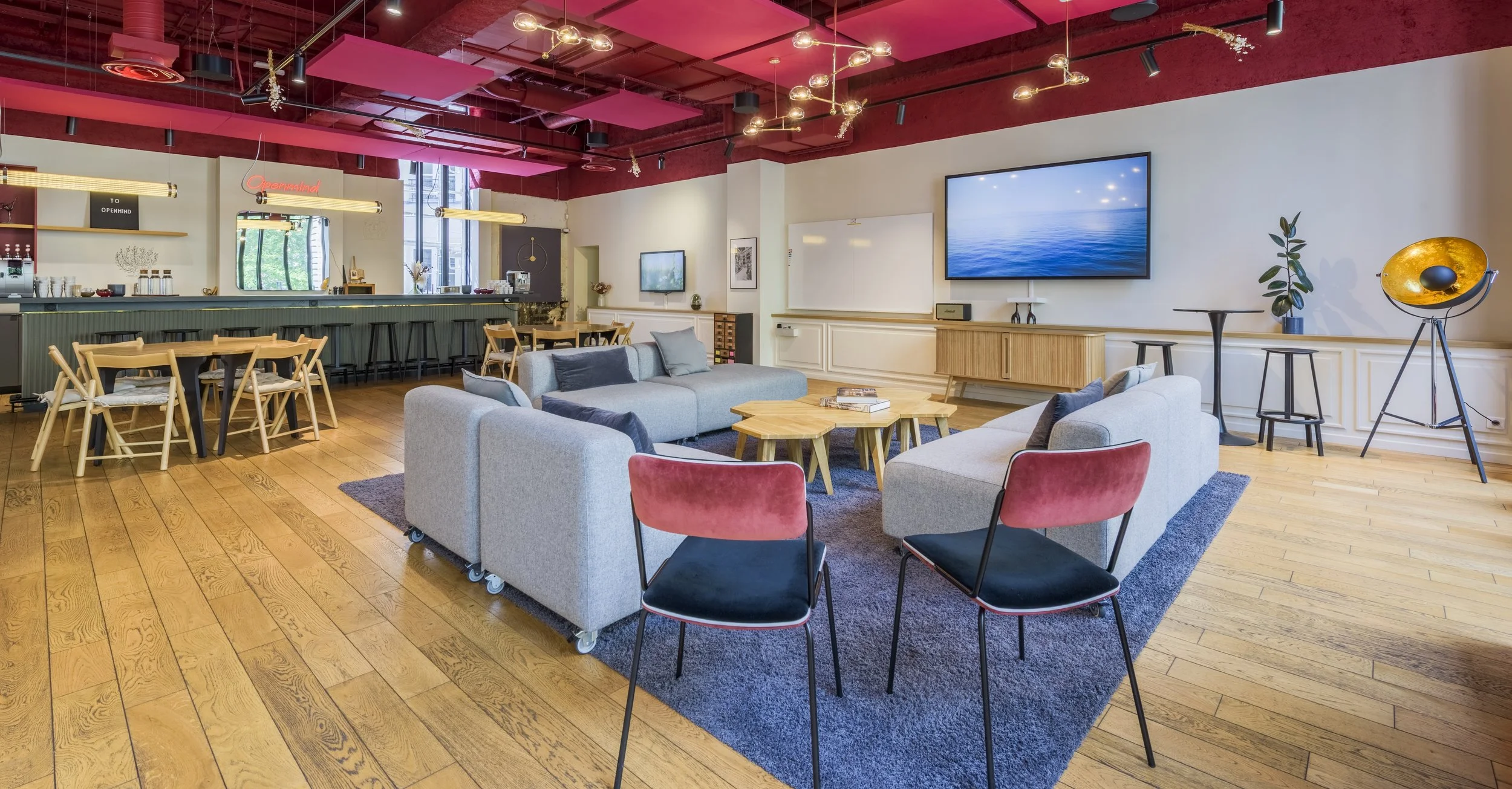Intérieur d'un salon moderne avec fauteuils, canapé, tables en bois, murs blancs, plafond rouge, et un espace bar. Mur blanc avec téléviseur, décor minimaliste et plantes.
