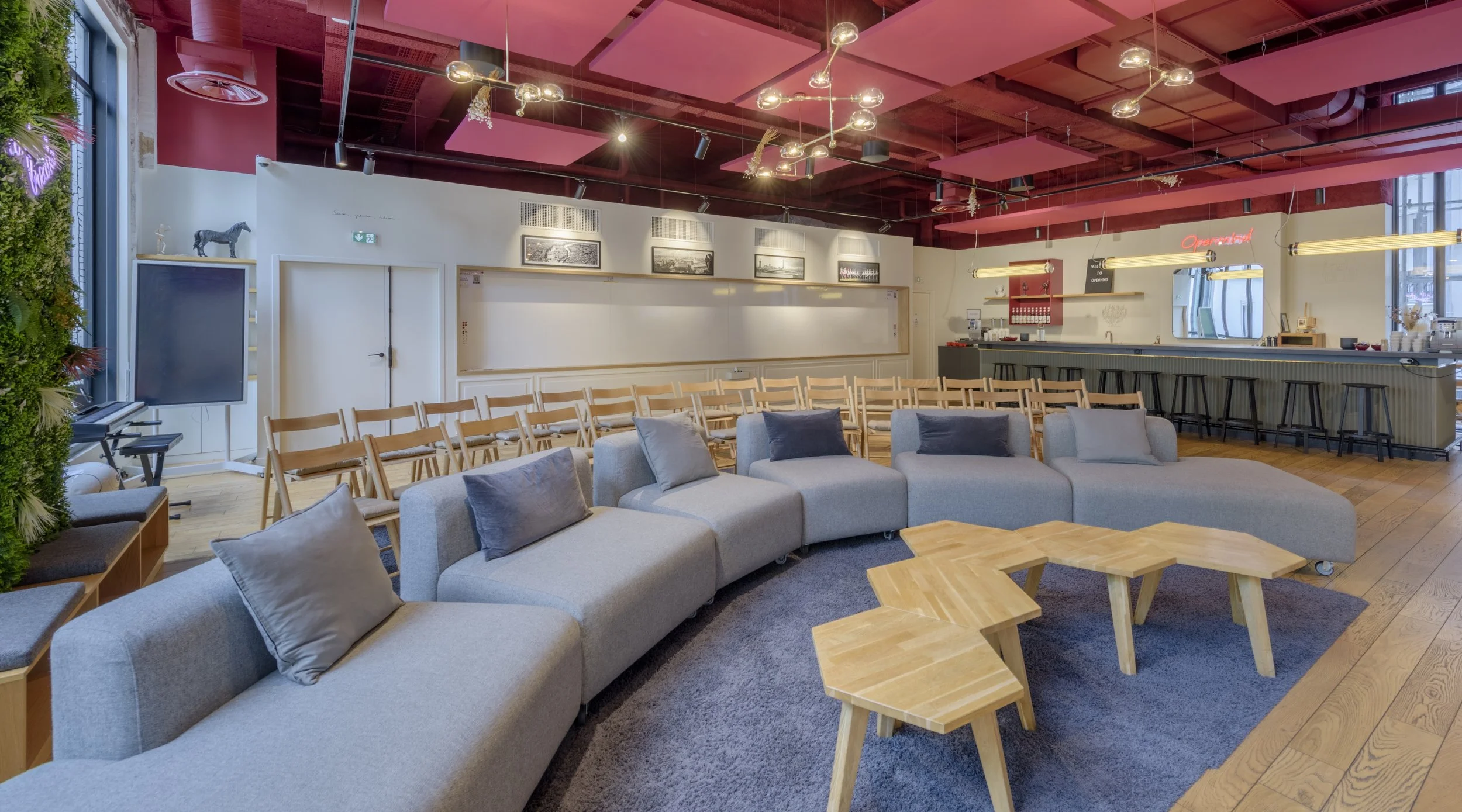 Intérieur d'un café ou bar moderne avec un grand canapé gris et tables en bois, bar en arrière-plan avec tabourets noirs, décoratif mur de plantes et lumineux plafonds roses.