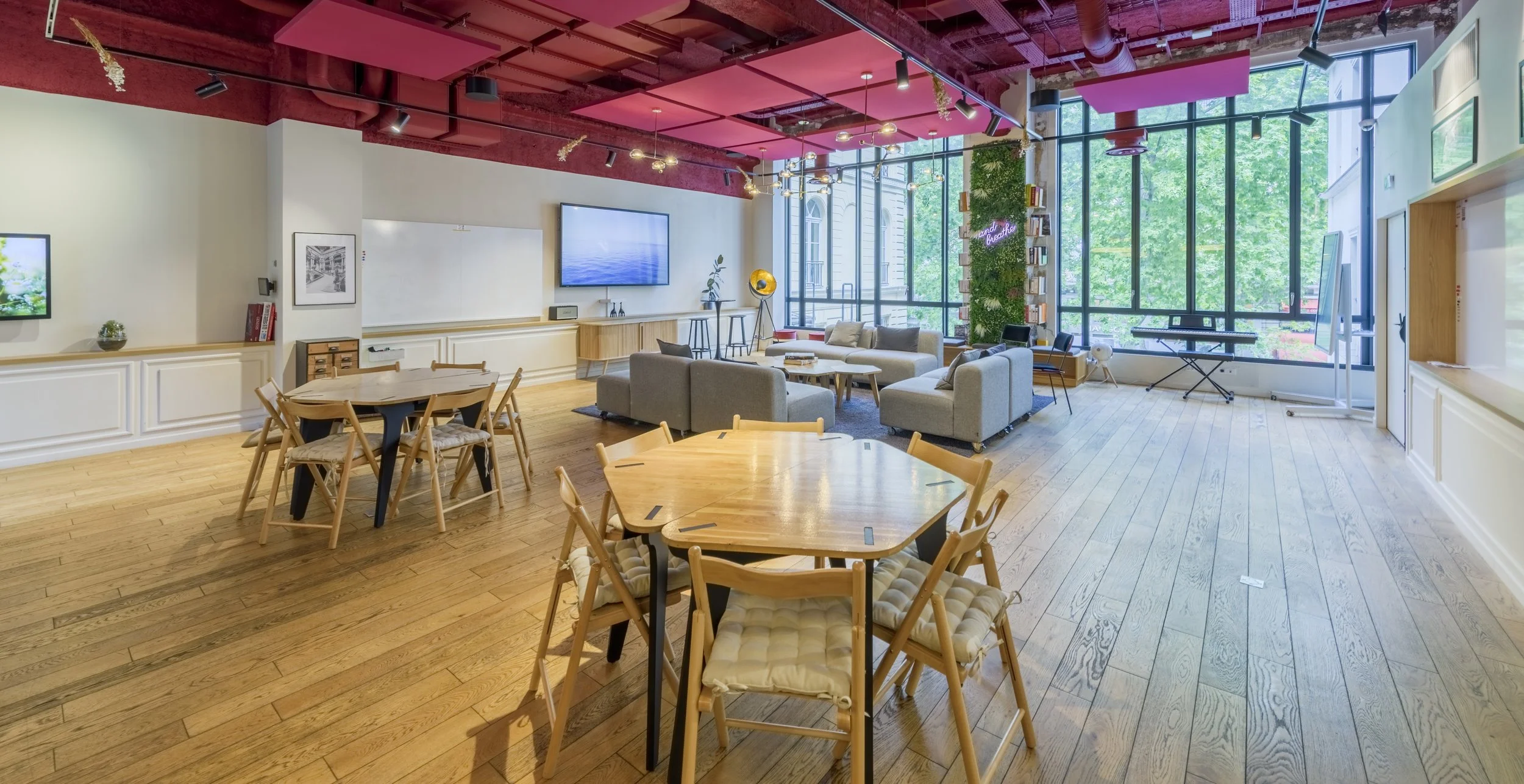 Espace de coworking ou de réunion lumineux avec grands fenêtres, mobilier en bois, canapés gris, tables en bois, et équipement audiovisuel.