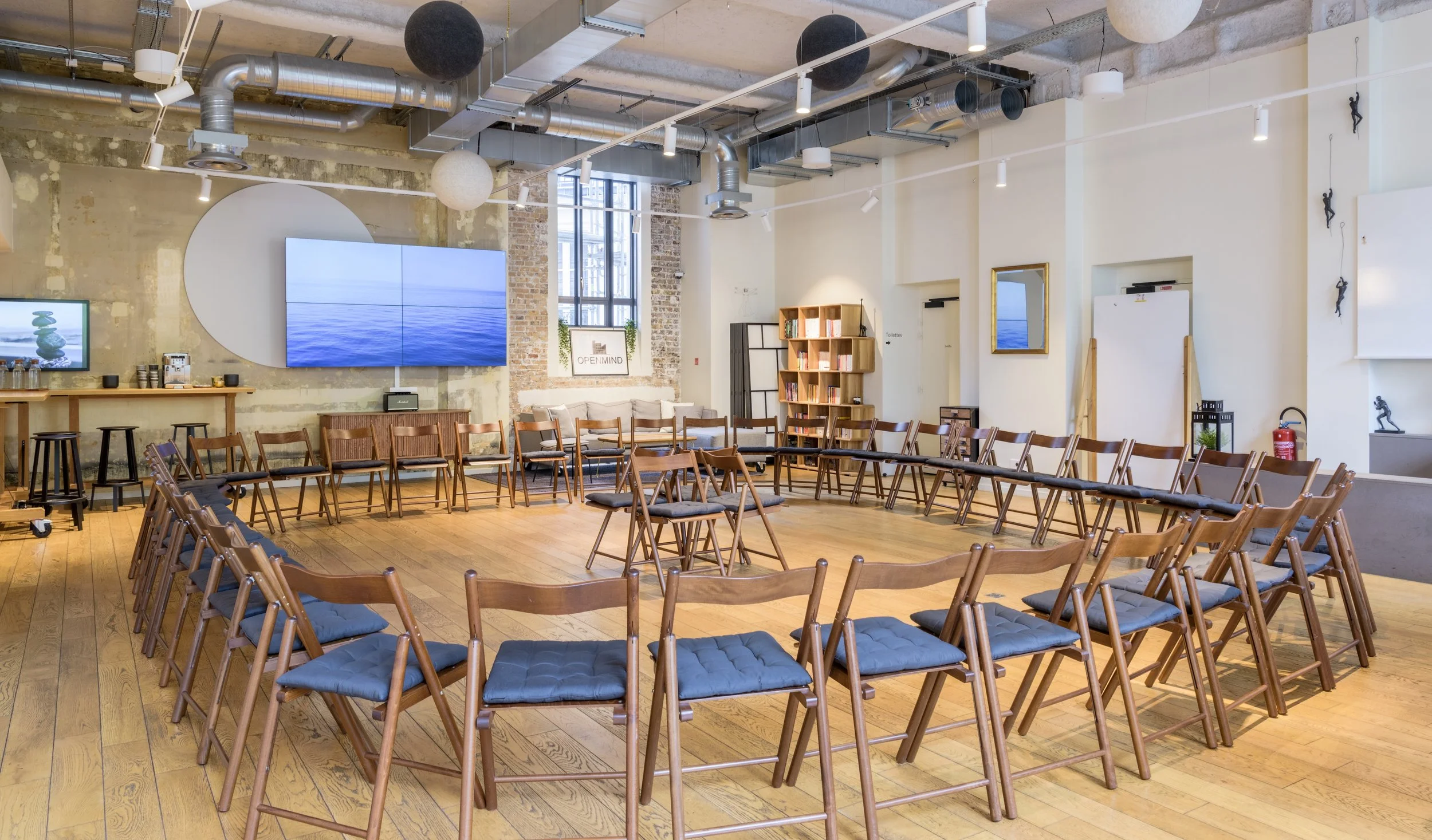 Salle de réunion avec plusieurs chaises en bois disposées en cercle, grande télévision à l'arrière, murs en briques et blanc, décor moderne avec des plantes et un miroir.