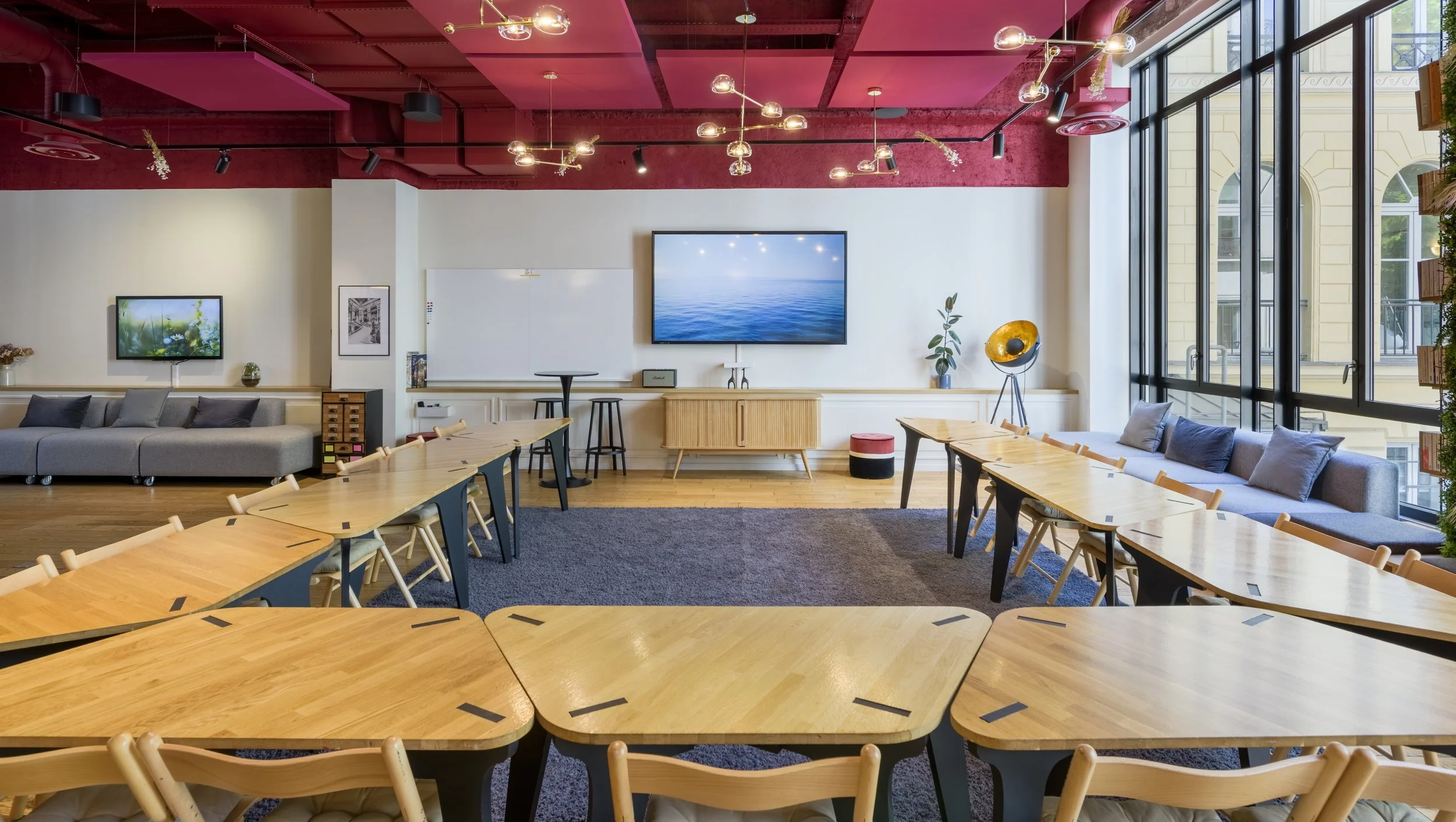 Salle de réunion ou espace de travail collaboratif avec des tables en bois disposées en U, un mur blanc avec deux écrans, une grande fenêtre laissant entrer la lumière naturelle, et une décoration moderne avec des lampes suspendues et des plantes.