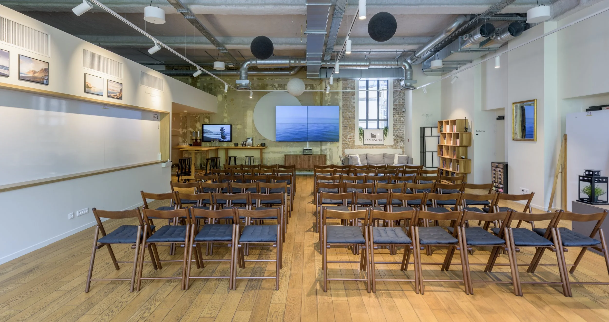 Une salle de conférence ou de présentation avec des rangées de chaises en bois, un mur blanc, un écran vidéo mural, et un espace bar en arrière-plan.