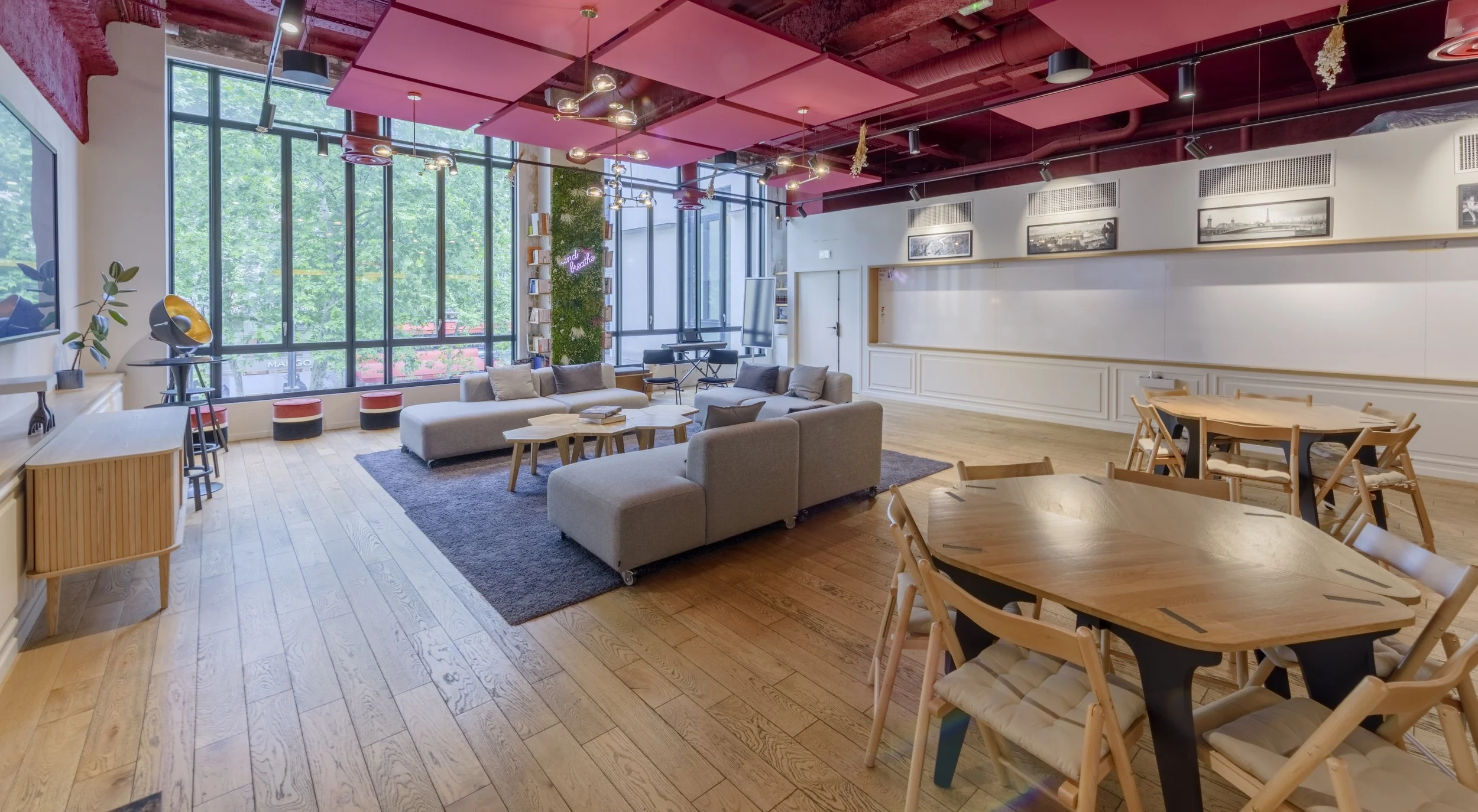 Intérieur moderne d'un espace de coworking ou salon avec grandes fenêtres, sofas gris, tables rondes en bois, et décoration contemporaine.