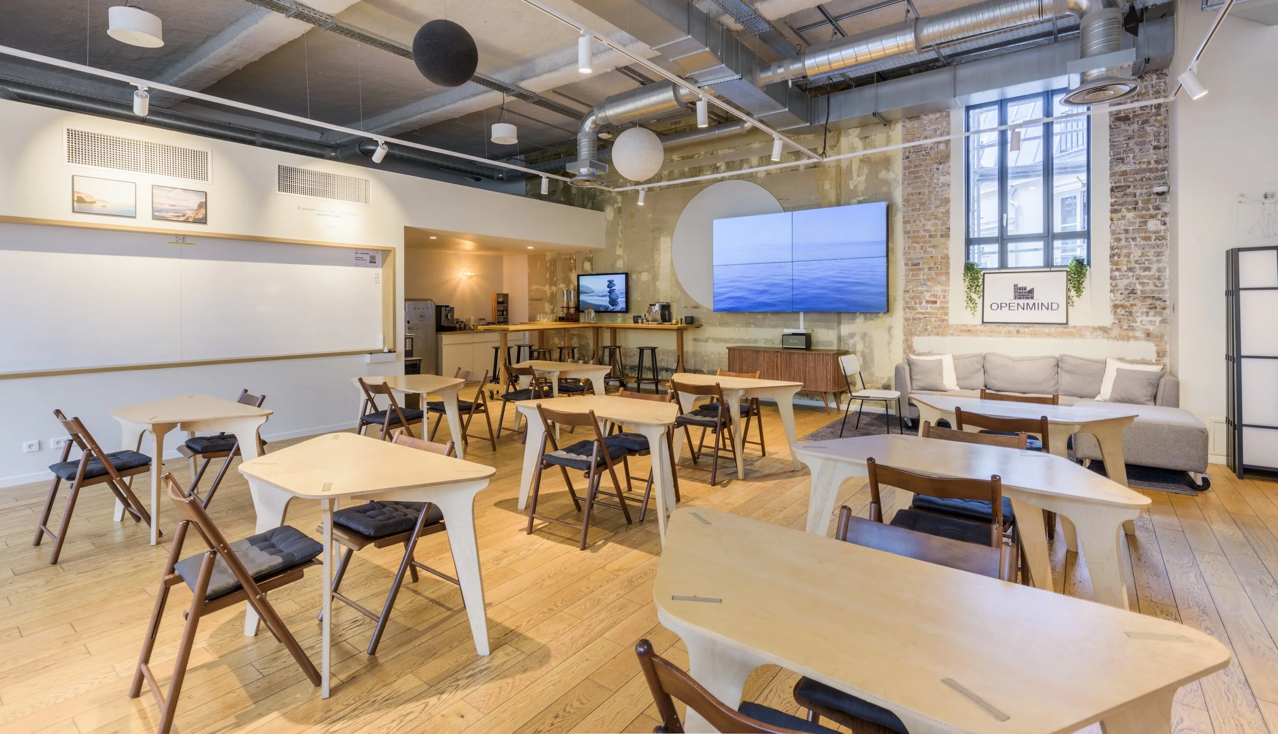 Salle de réunion ou espace de coworking moderne avec plusieurs tables en bois, chaises empilables, grand écran mural, sofa, et décor industriel avec mur en briques, plafond exposé et luminaires suspendus.