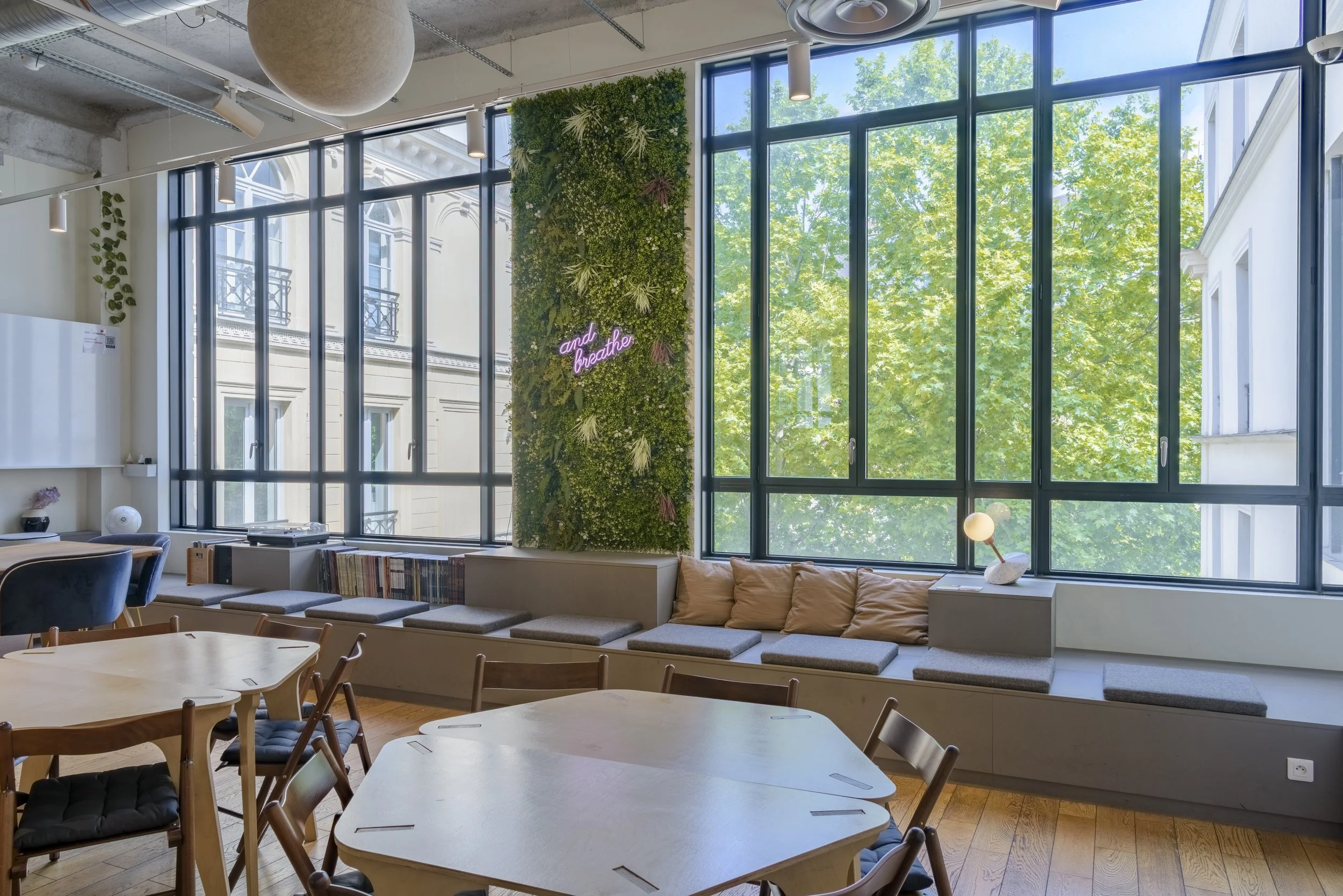 Intérieur lumineux avec grande baie vitrée, murs en béton, espace de détente avec coussins, mobilier en bois, mur végétal avec néon rose "and breathe"