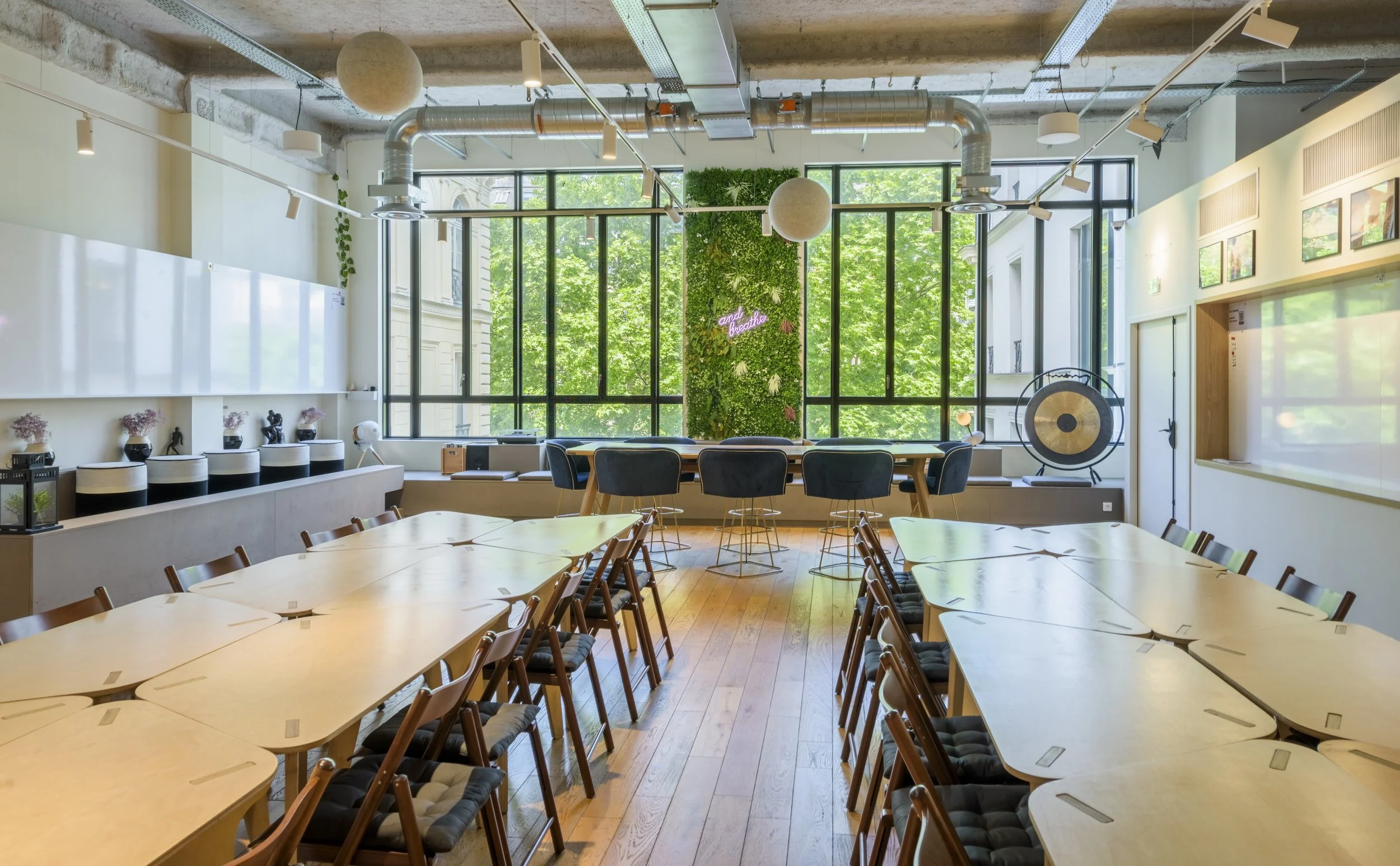 Salle de réunion lumineuse avec grandes fenêtres, tables en forme de U, chaises autour, décoration verte avec un panneau en mur végétal, instruments de musique et affichage de dessins