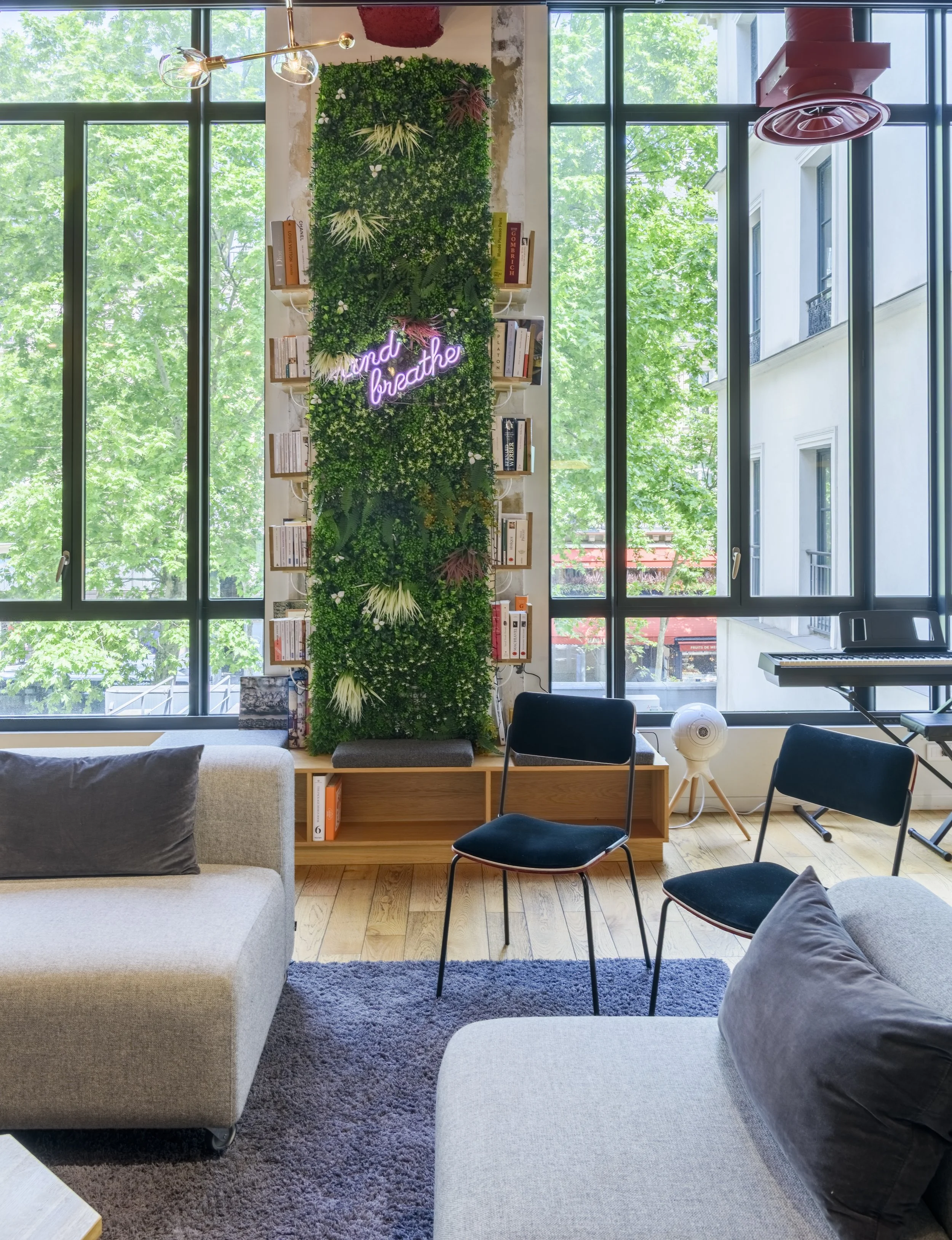 Salon avec grands fenêtres, mur végétal avec néon 'find breath', livres, piano, chaises, canapé, tapis, et un ventilateur.