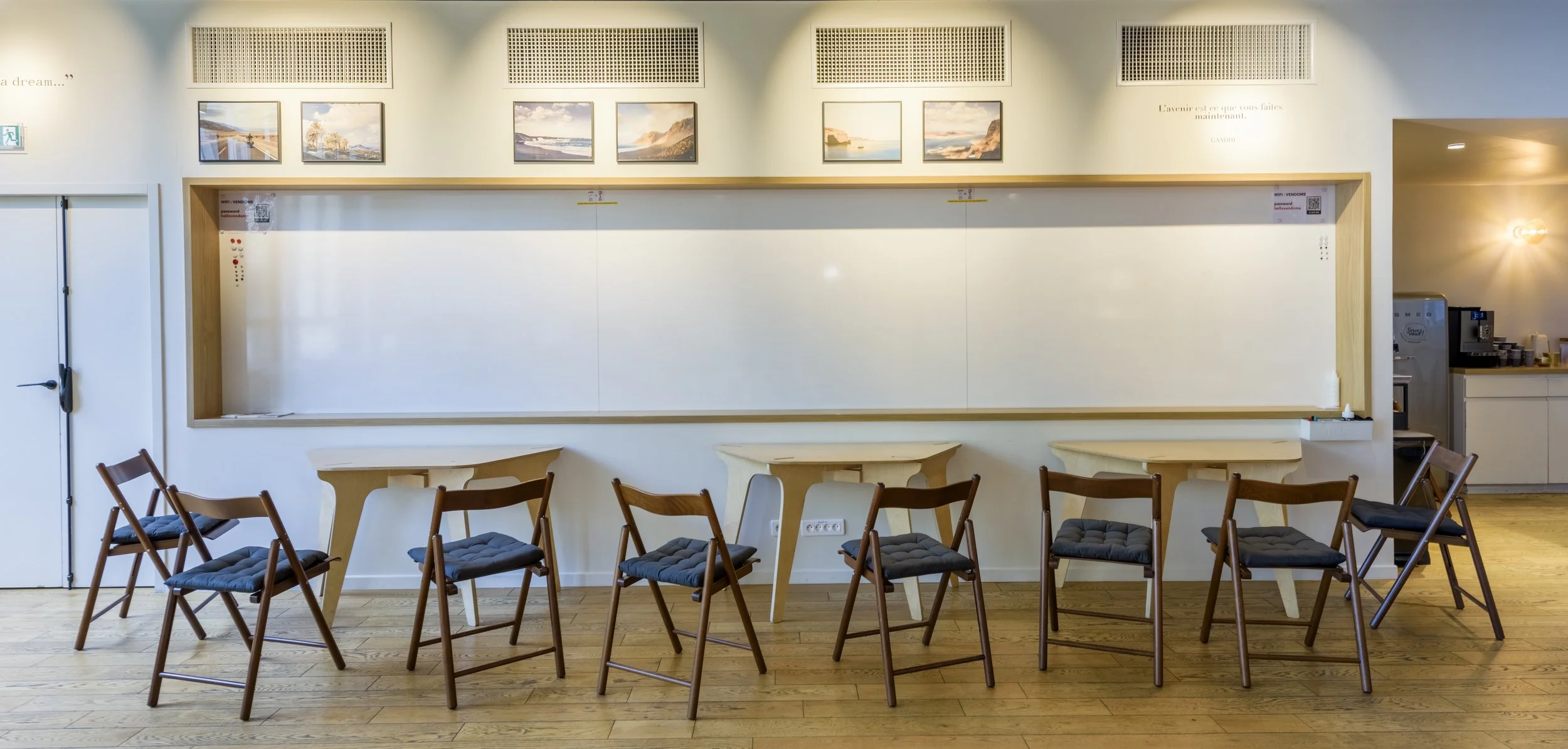 Salle de réunion vide avec huit chaises en bois avec coussins noirs, trois petites tables blanches, tableau blanc vide, affiches de paysages au-dessus, et coin café en arrière.