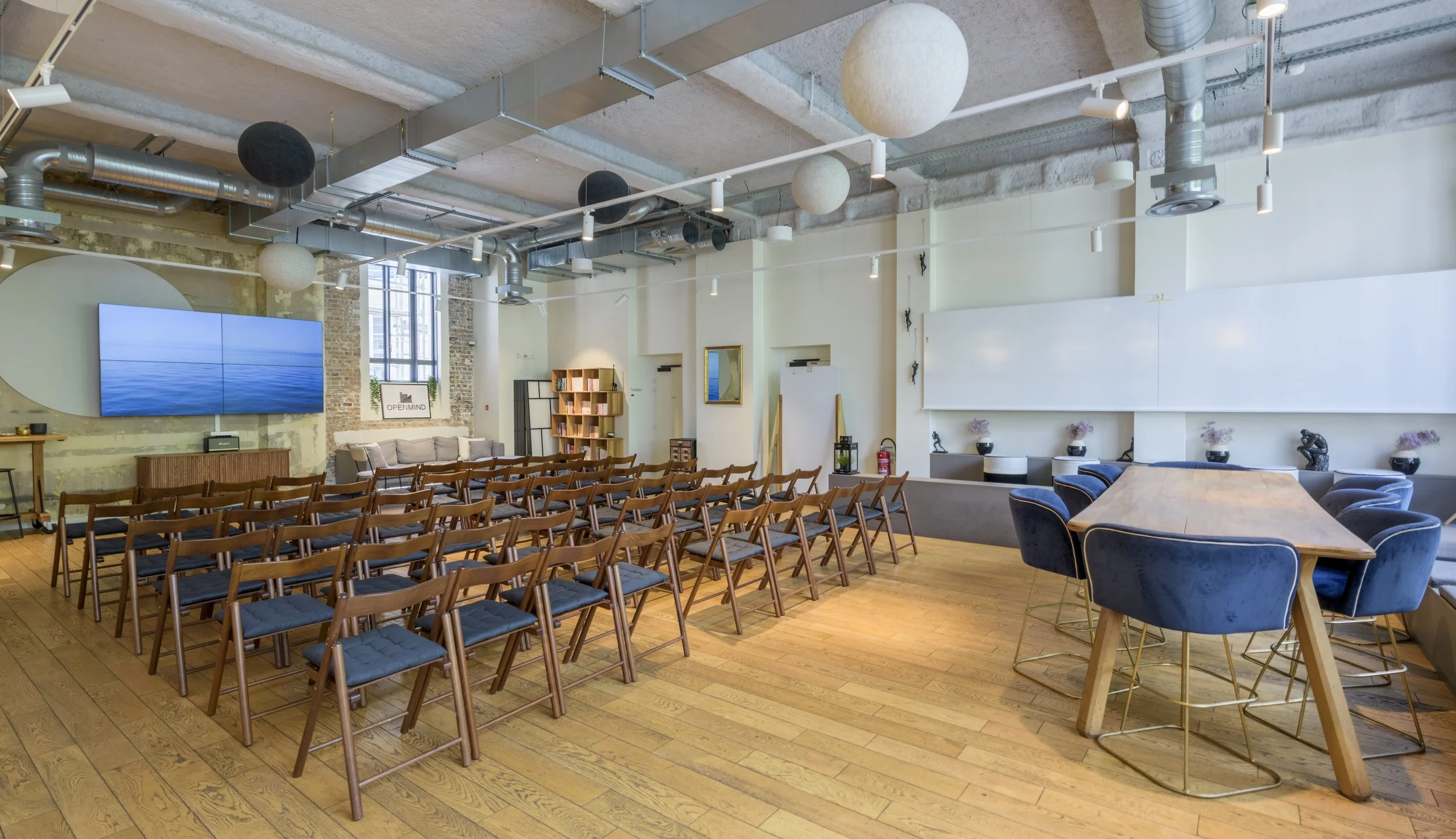 Une salle de conférence ou de réunion moderne avec des chaises en bois disposées en rangées, un grand écran à écran plat affichant une image de l'océan, une table en bois avec des chaises bleues, et diverses décorations dont des livres, des tableaux 