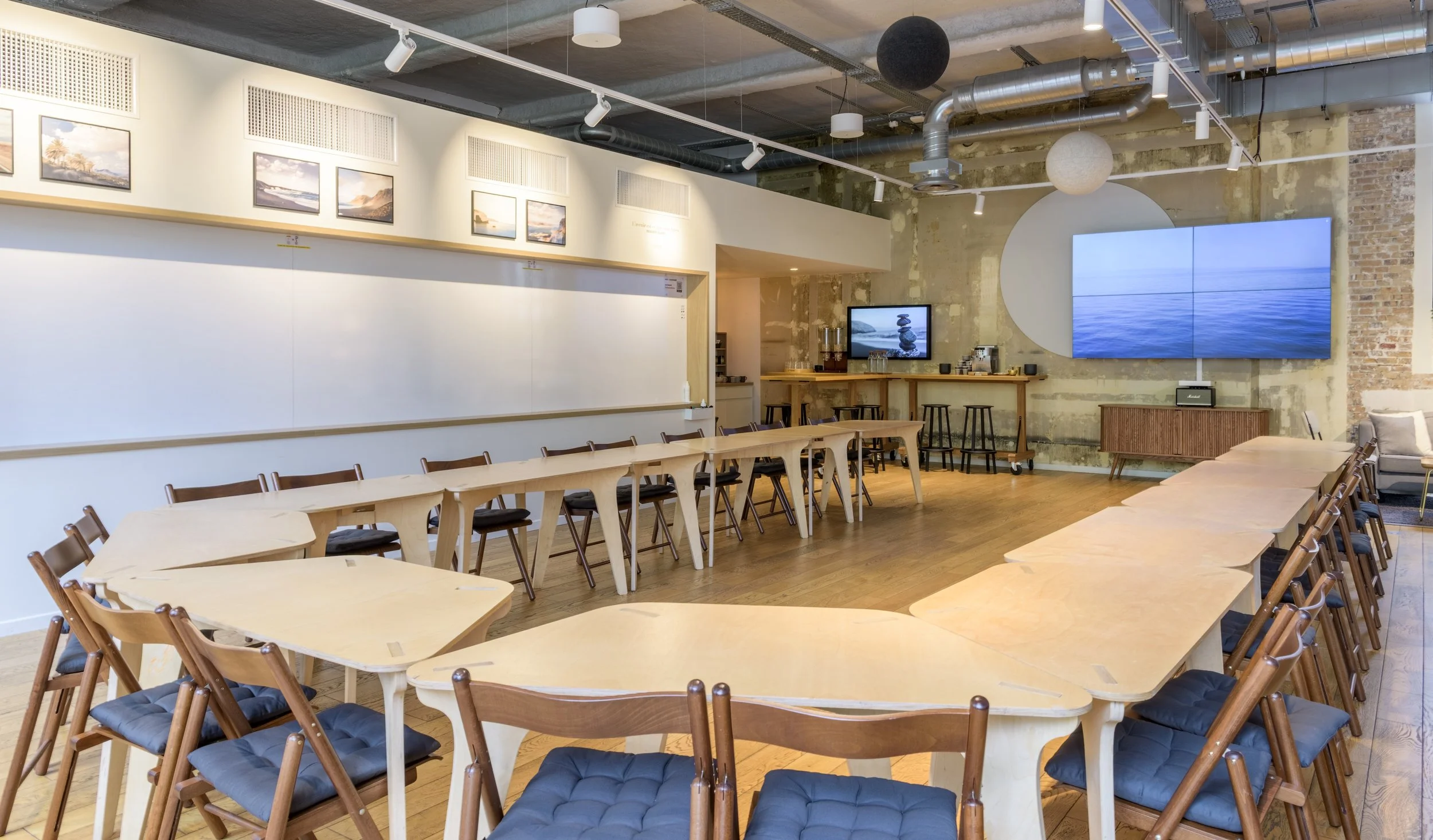 Une salle de réunion ou un espace de coworking avec une grande table en forme de U, des chaises en bois avec des coussins noirs, des écrans muraux affichant une scène maritime, et un bar avec des sièges au fond. Décor moderne avec un plafond industri