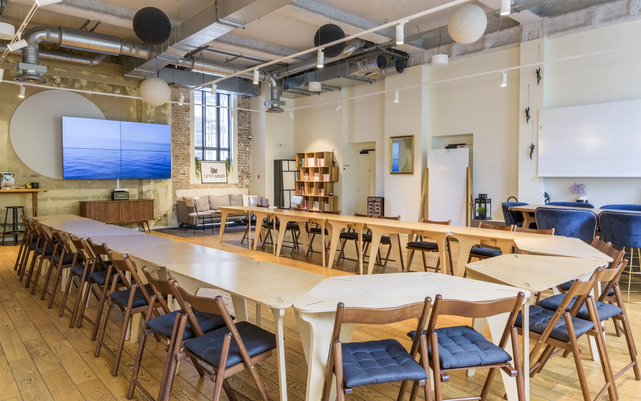 Salle de réunion avec tables en forme de U, chaises en bois avec assises en tissu noir, écran plat sur un mur en brique, ambiance lumineuse avec lumières suspendues et décor moderne.
