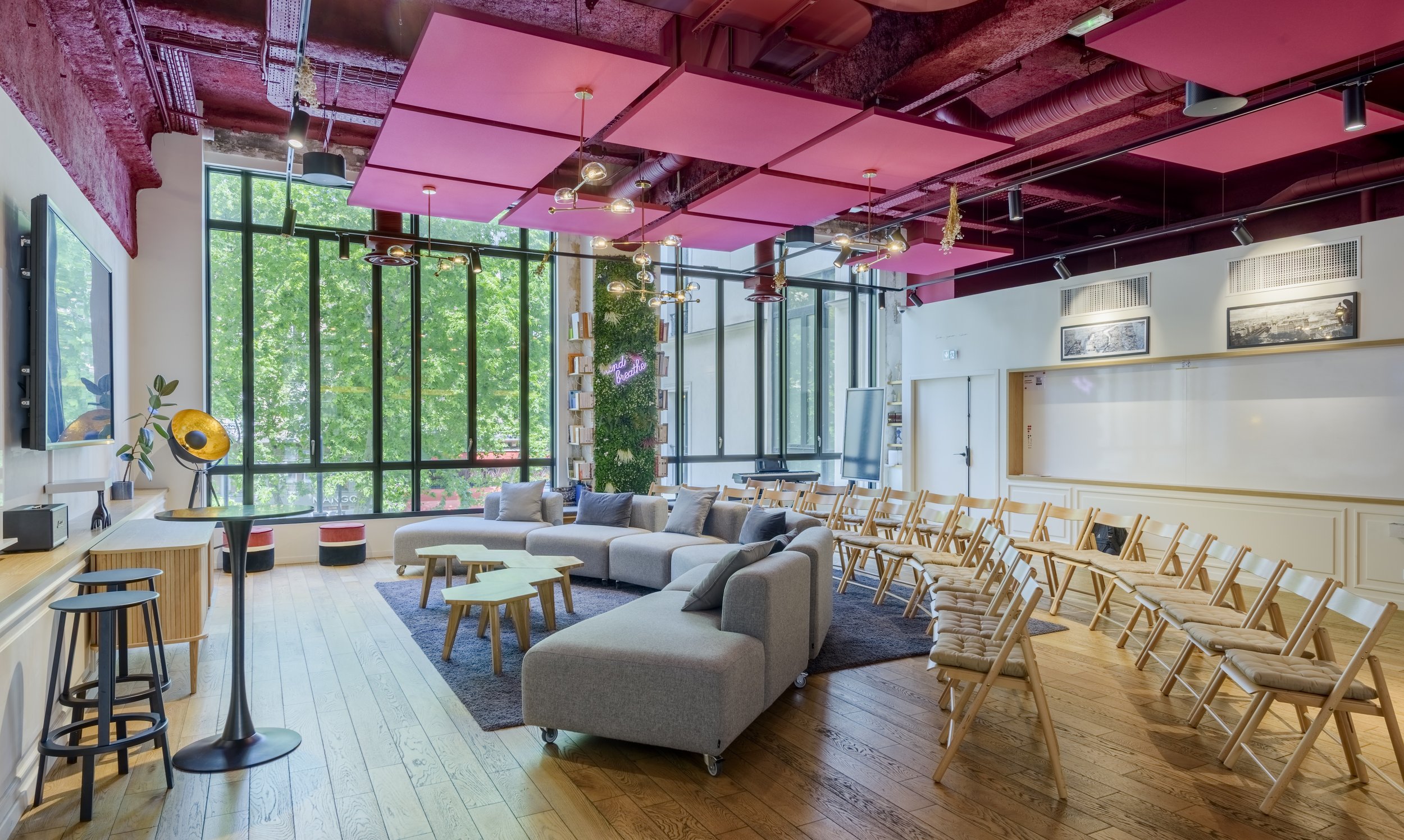 Salon moderne avec grands fenêtres, canapé gris, chaises en bois, tables basses, décoration éclectique, plafond coloré, et écrans