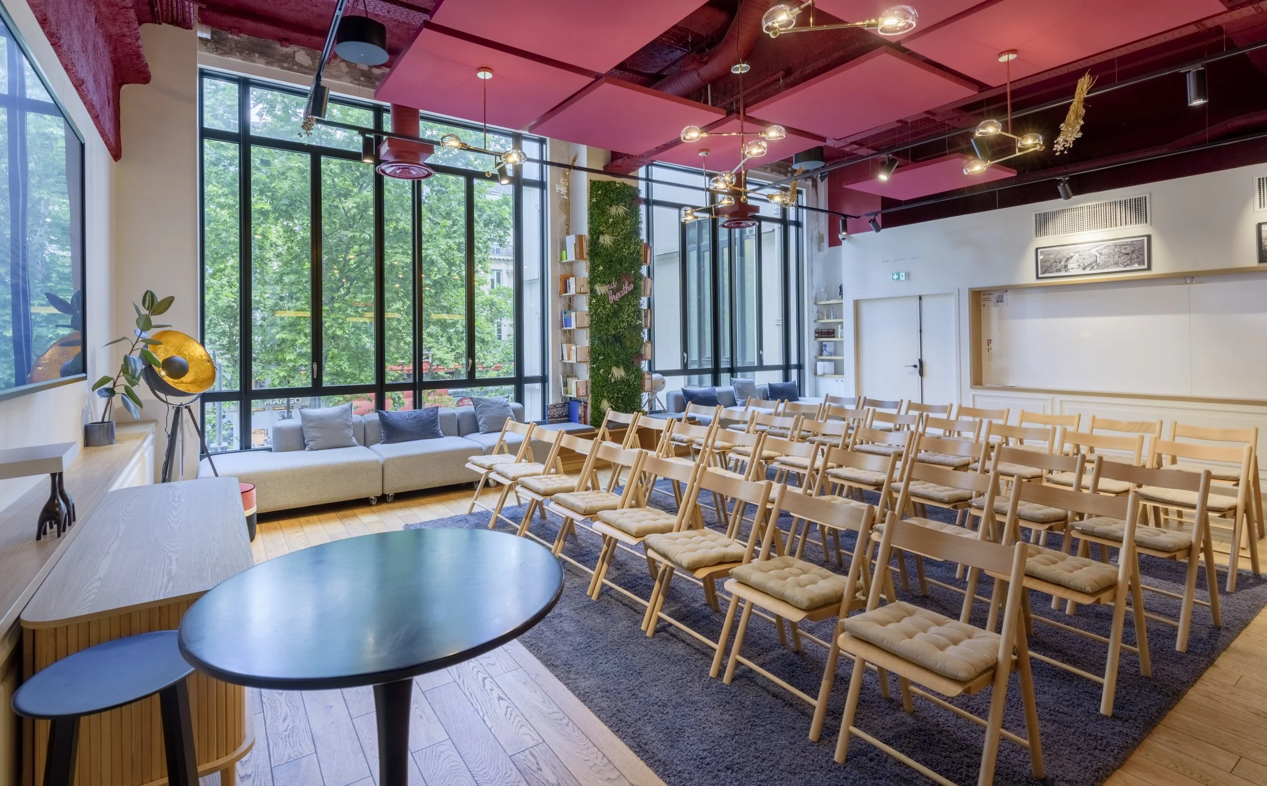 Salle de conférence moderne avec des rangées de chaises en bois, un canapé gris près des grandes fenêtres, et des décorations élégantes au plafond