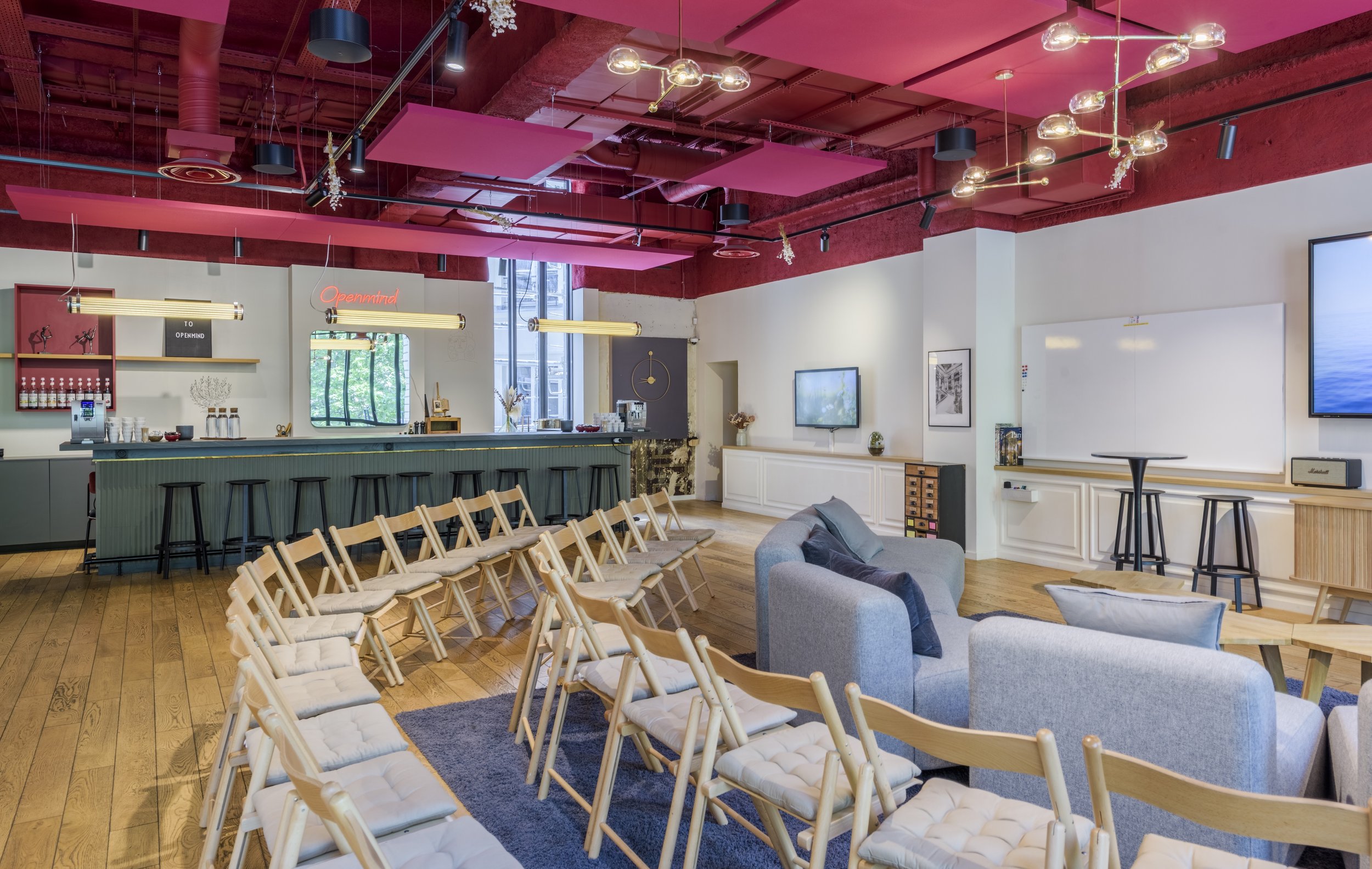 Interior design d'un espace de coworking ou salon de réunion avec des fauteuils, chaises en bois, un canapé gris, une table en bois, une kitchenette au fond, et un tableau blanc, éclairage moderne, plafond rose et murs blancs.