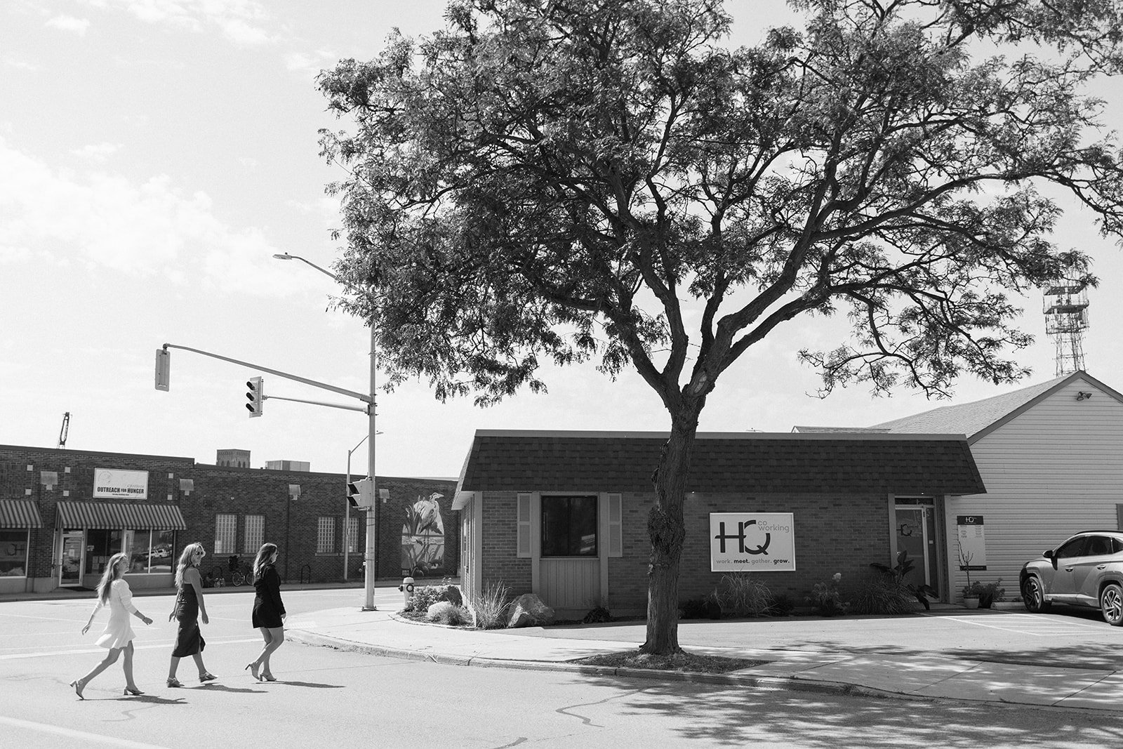 A black and white photo of a tree in front of a building with a sign that reads "HQ" and slogan "work. meet. gather. grow." Three women walk across the street, with traffic lights and a building with a mural in the background.