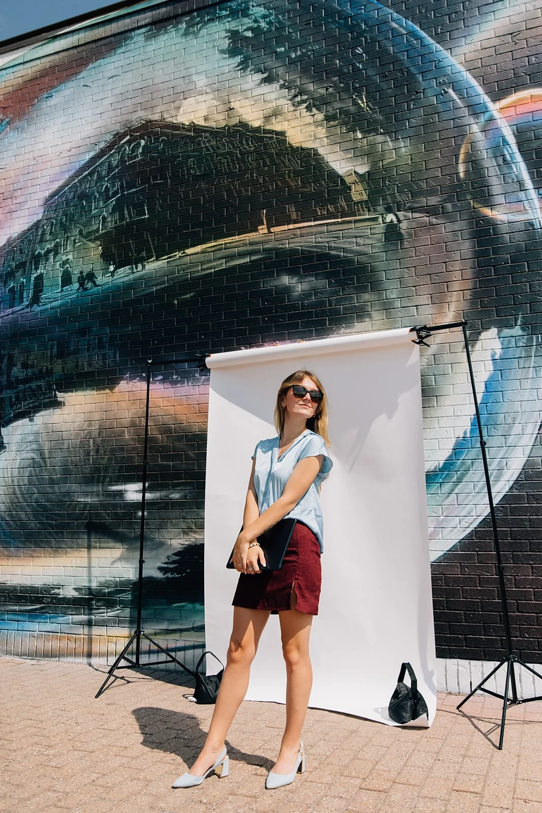 A woman standing outdoors in front of a graffiti mural on a brick wall, with a white backdrop and photography equipment around her.