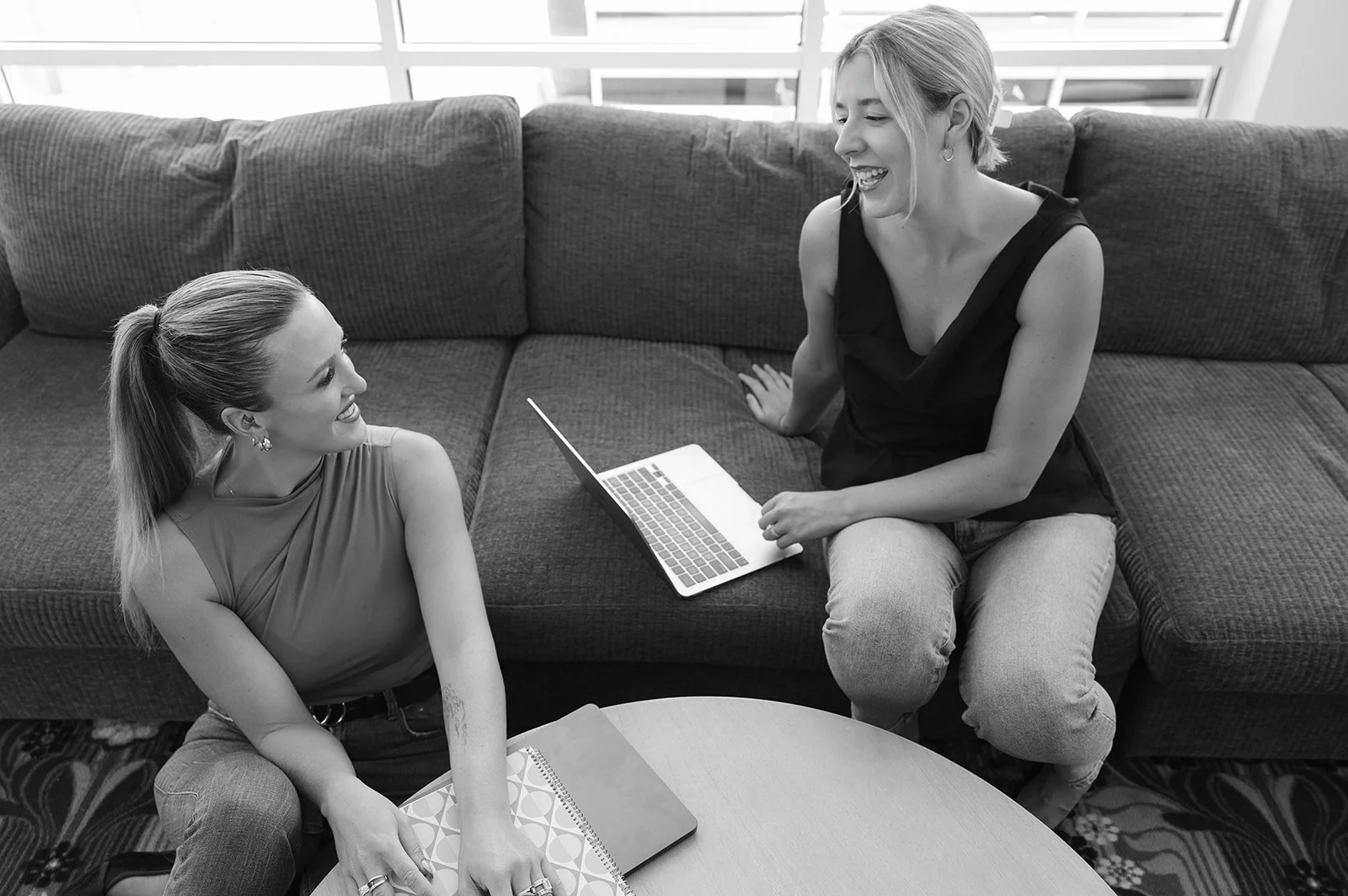 Two women smiling and talking in a living room, one sitting on a sofa with a laptop on her lap and the other sitting on a chair with notebooks in front of her.