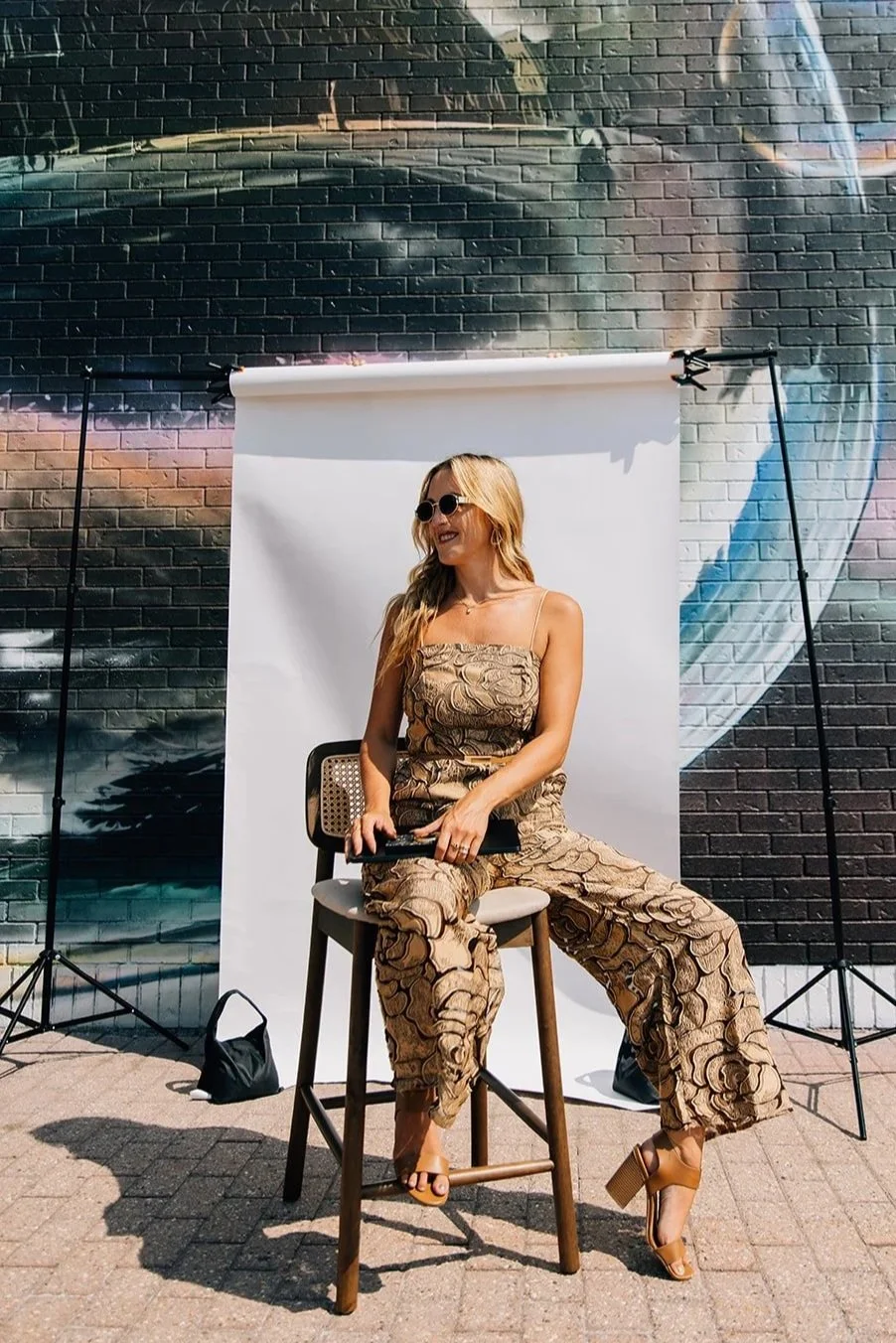 A woman in patterned beige jumpsuit sitting on a wooden stool in front of a white backdrop, with a graffiti-covered brick wall in the background. She is wearing sunglasses, beige high-heeled sandals, and has a black clutch in her hand.