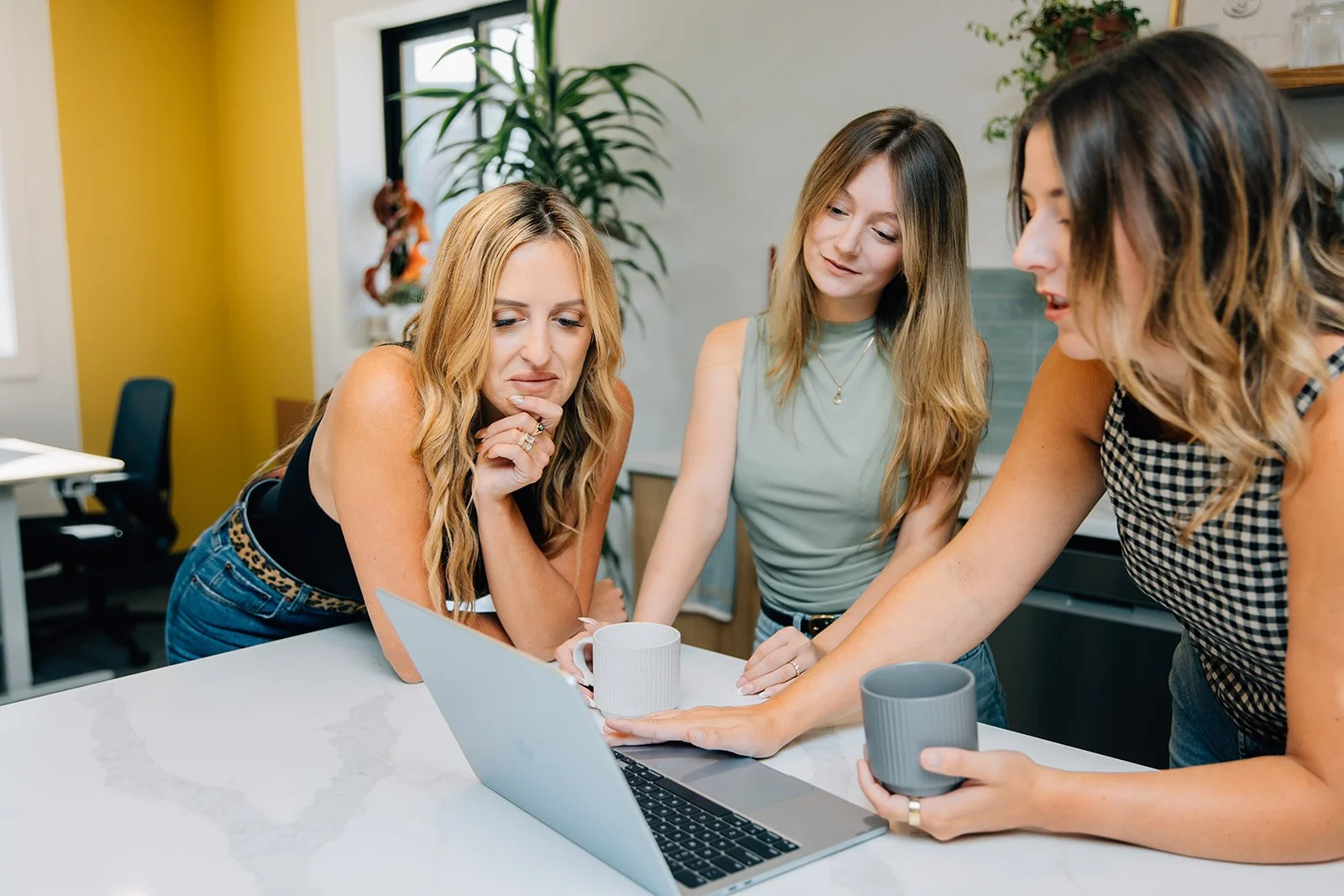 Three women gathered around a laptop in a modern office, looking at the screen with interest. They are holding coffee mugs and appear to be discussing something on the laptop.
