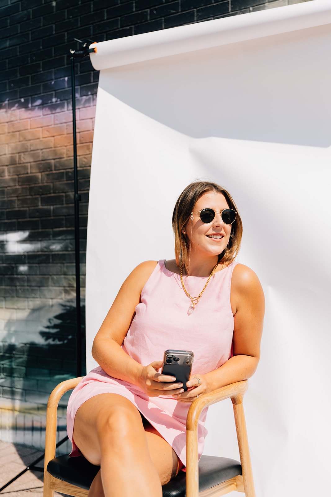 A woman wearing sunglasses and a pink sleeveless dress, sitting on a wooden chair outside, holding a smartphone, with a white backdrop and dark brick wall in the background.