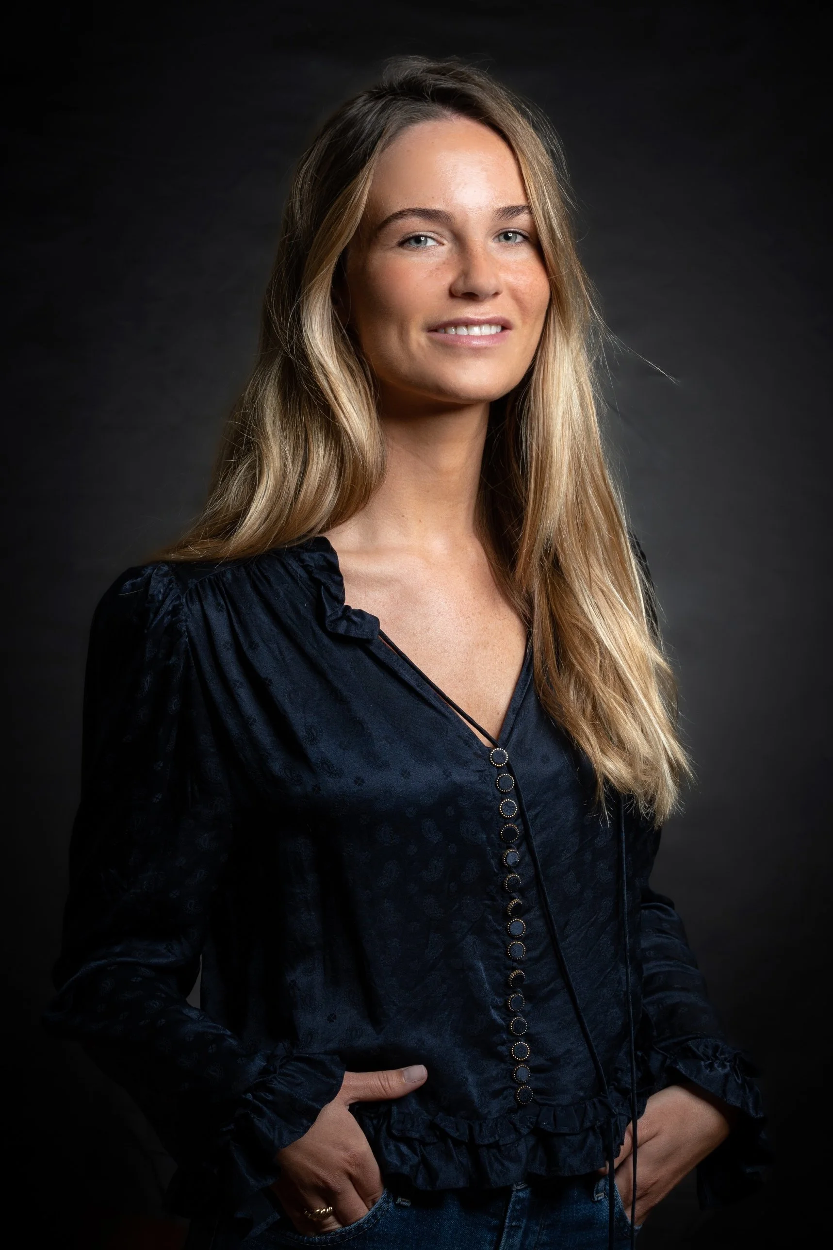 Jeune femme aux cheveux longs et blonds, portant une blouse noire avec des détails en cercle, positionnée de profil contre un fond sombre.