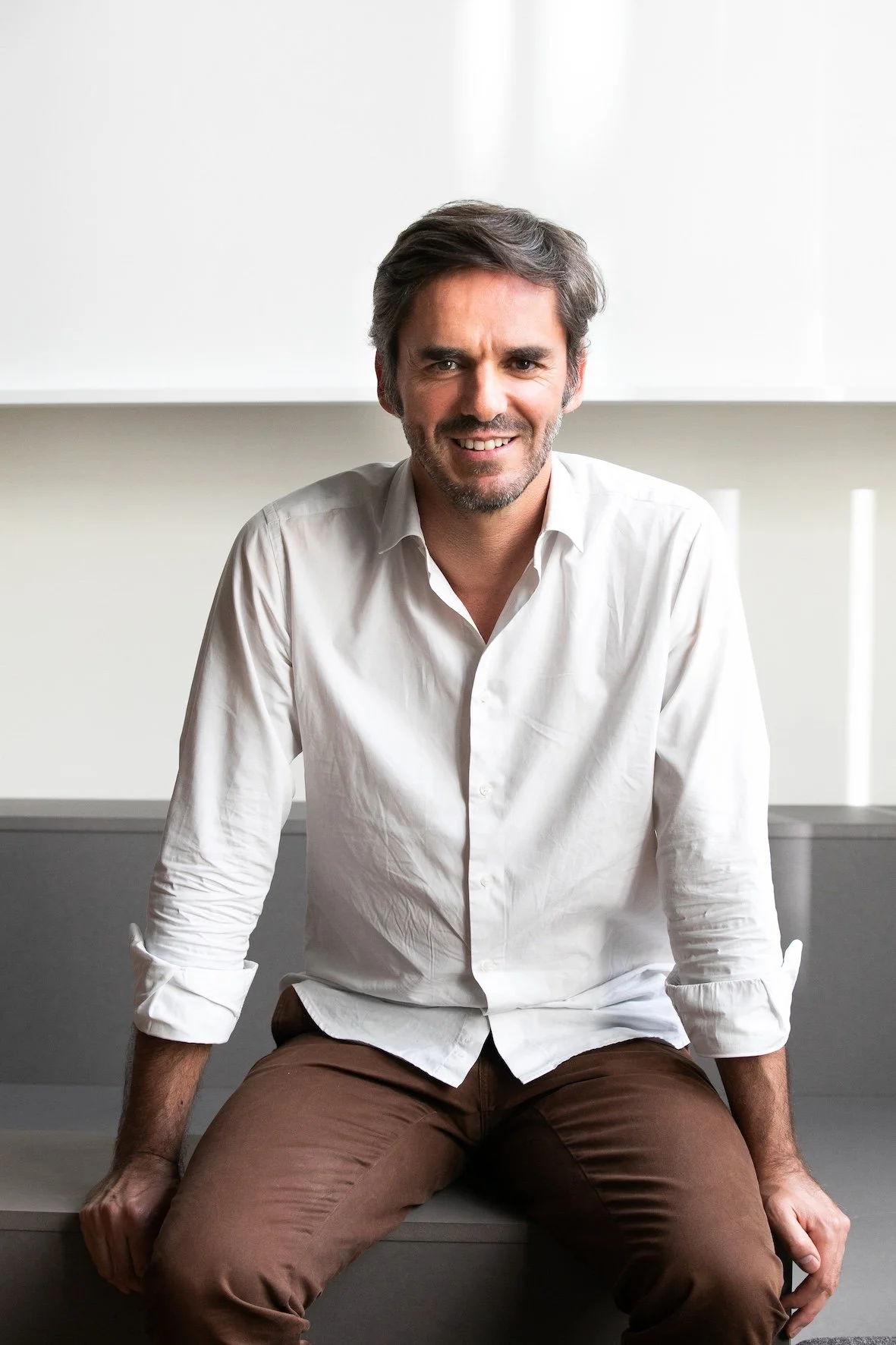 Homme souriant en chemise blanche et pantalon marron, assis sur une surface grise dans un environnement lumineux et minimaliste.