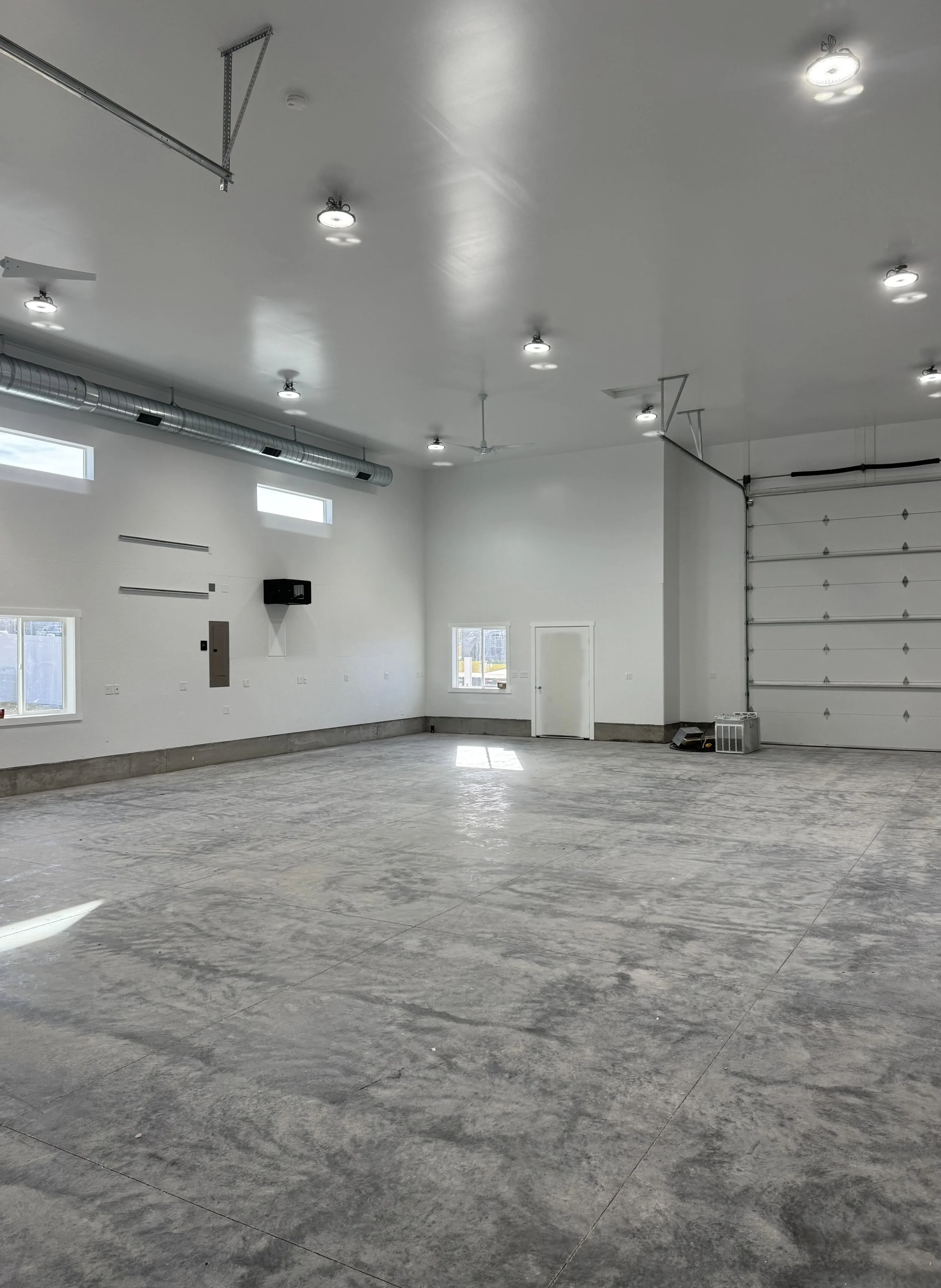 Empty commercial or industrial garage or warehouse with concrete floors, white walls, high ceiling, windows, and a large garage door.