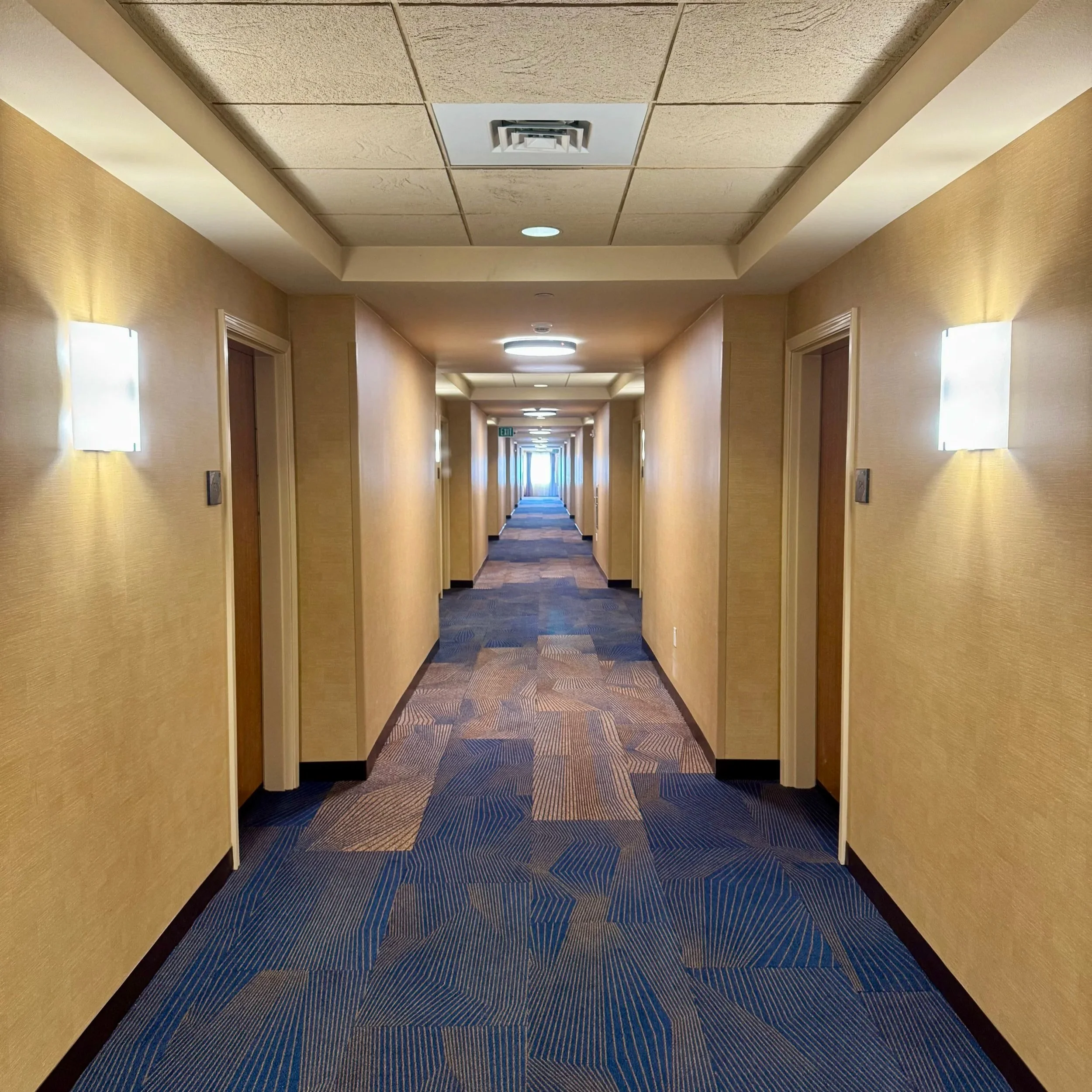 Empty hotel hallway with beige walls, blue patterned carpet, and wall-mounted lights, leading to a distant door at the end.